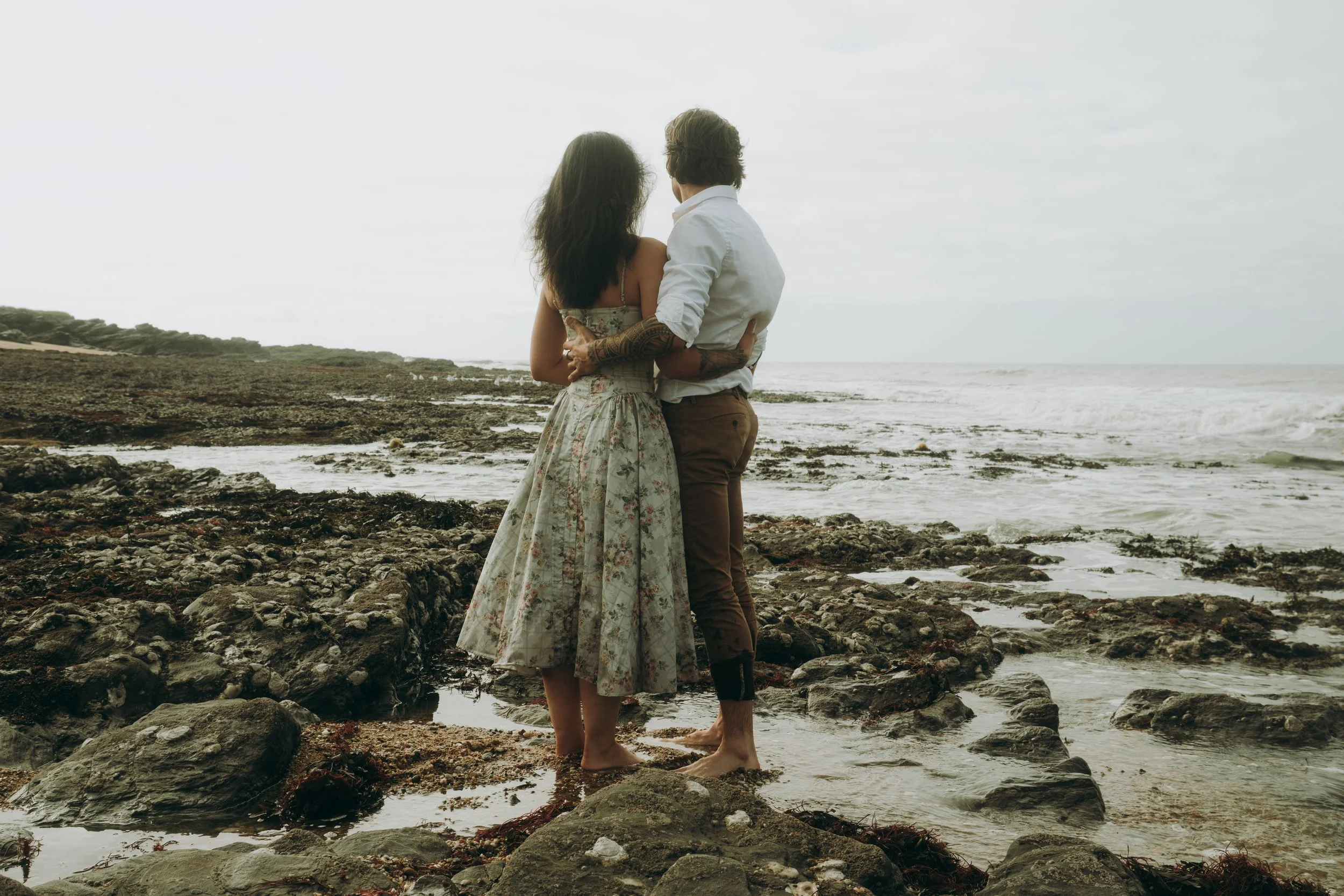 Un couple se tenant par la taille sur une plage rocheuse, regardant vers la mer sous un ciel nuageux.