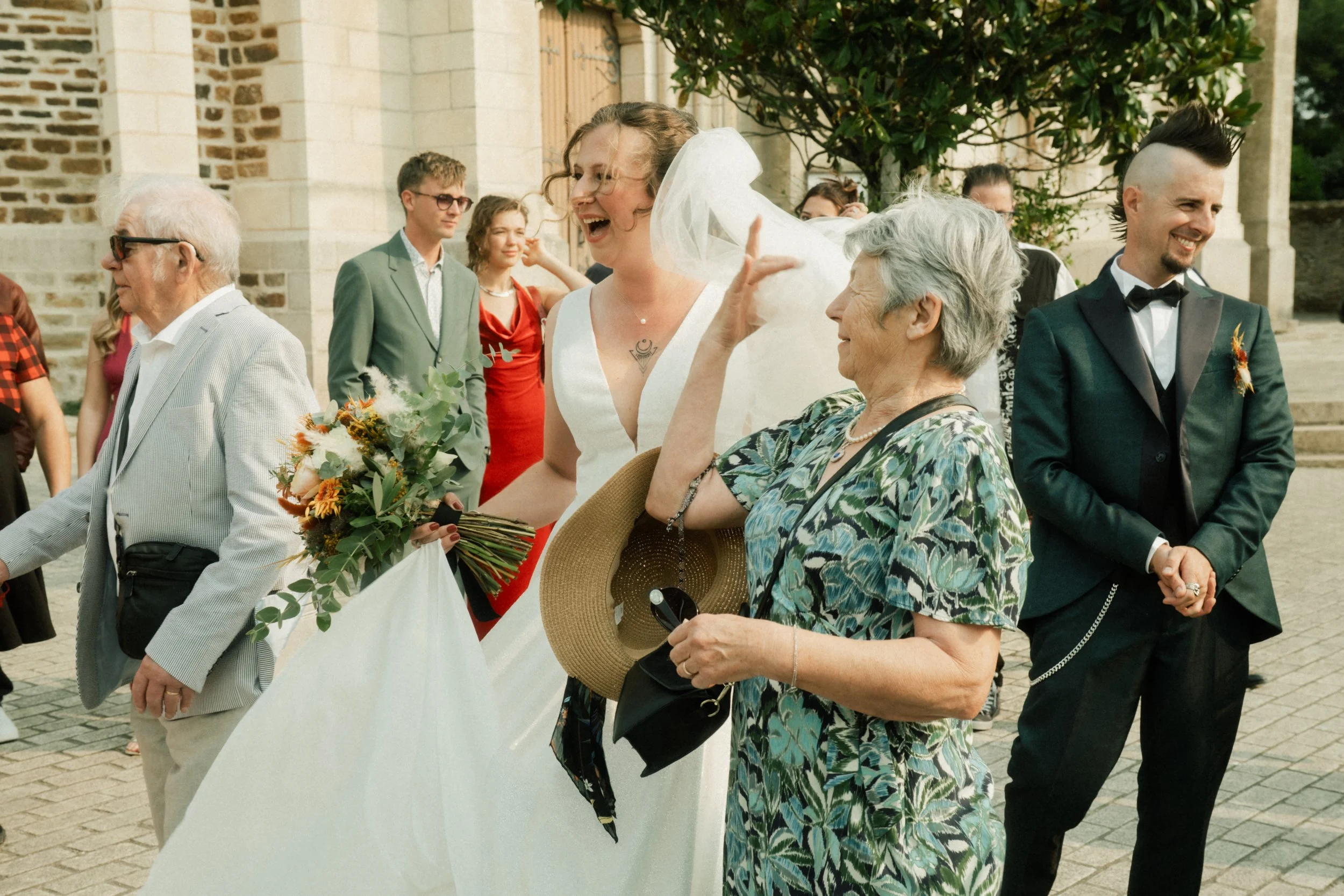 Un groupe de personnes habillées pour un mariage, avec une femme en robe blanche, souriante, entourée d'autres invités, certains souriants, certains tenant des fleurs ou des objets, devant une bâtisse en pierre.
