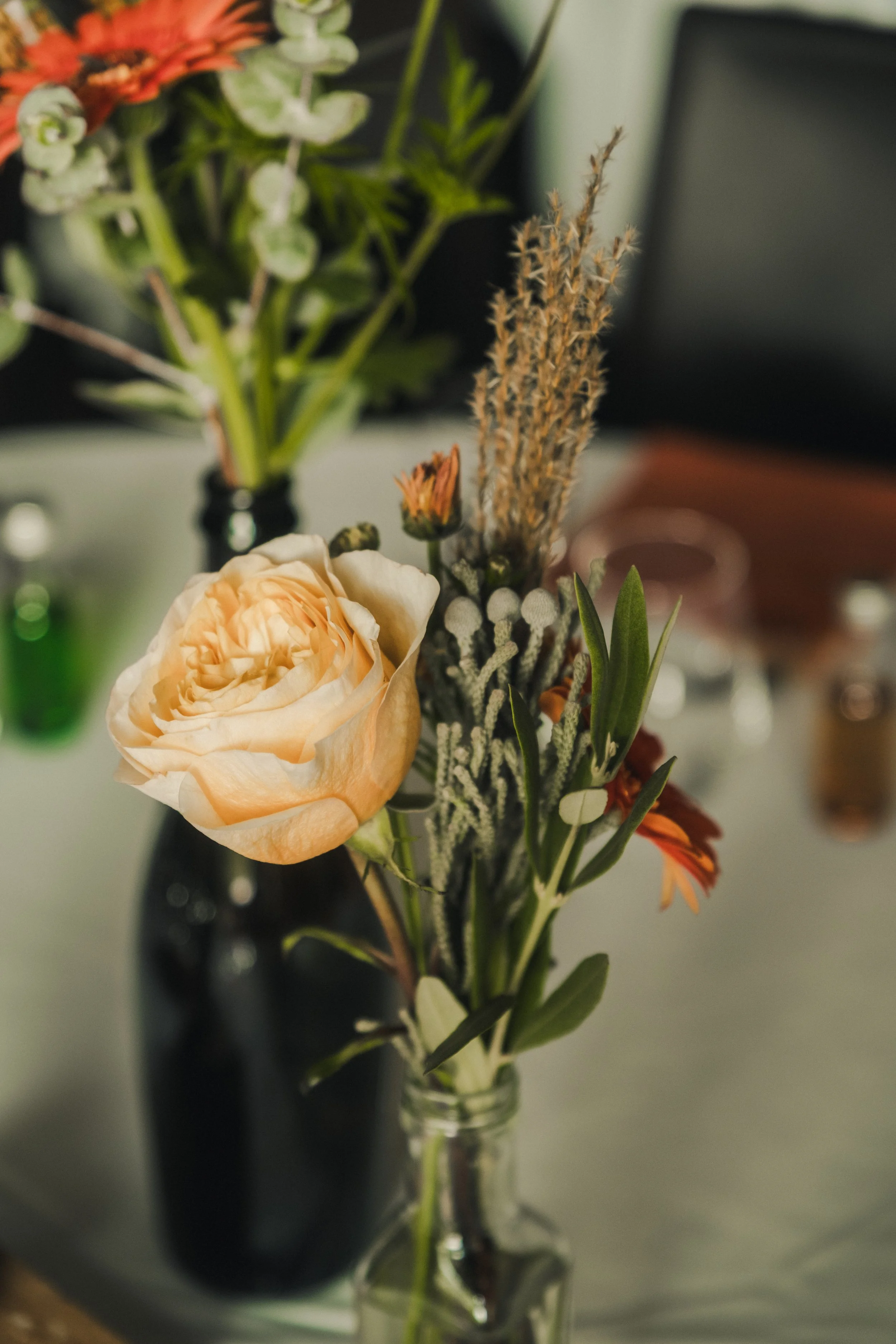 Un bouquet de fleurs dans un vase en verre, avec une rose beige, des fleurs orange, des fleurs blanches et des plantes vertes, en arrière-plan un autre bouquet dans un vase noir.