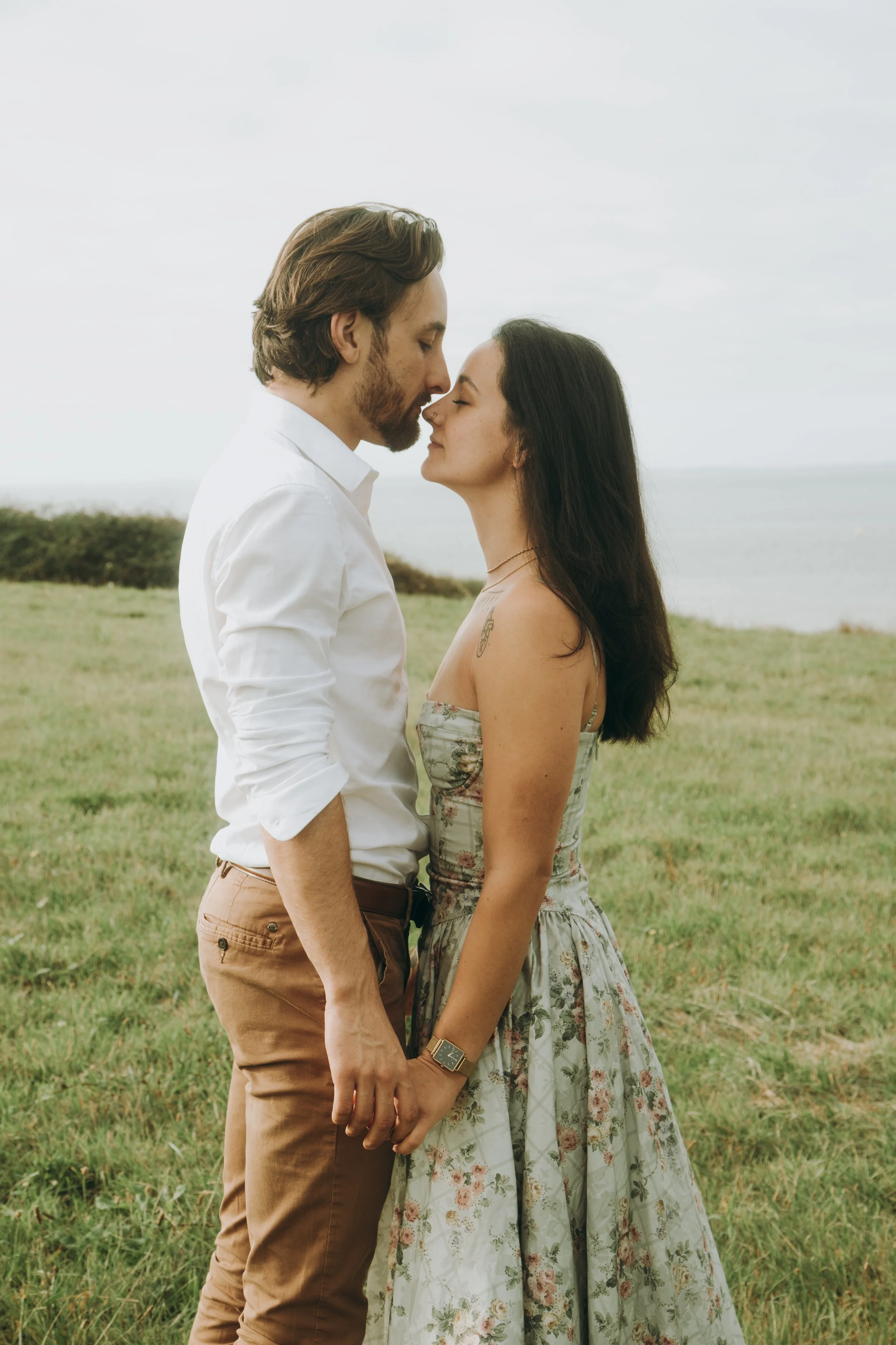 Un couple tente une embrassade, ils se tiennent la main et se regardent tendrement dans un parc près de la mer sous un ciel nuageux.