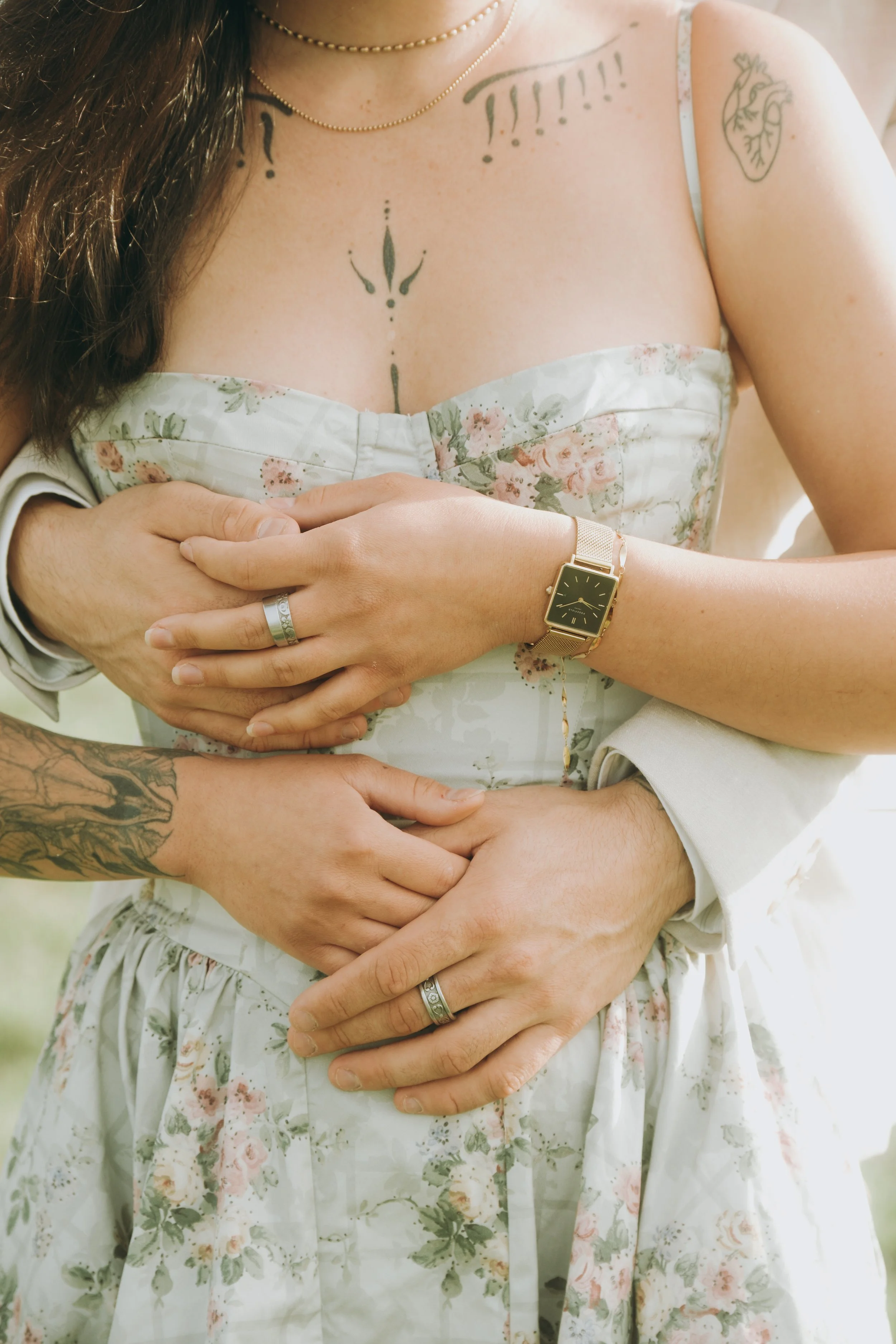 Gros plan des mains d'un couple marié, portant des alliances, avec une femme portant une montre dorée, une robe de mariage à motifs floraux, et un tatouage sur l'épaule.