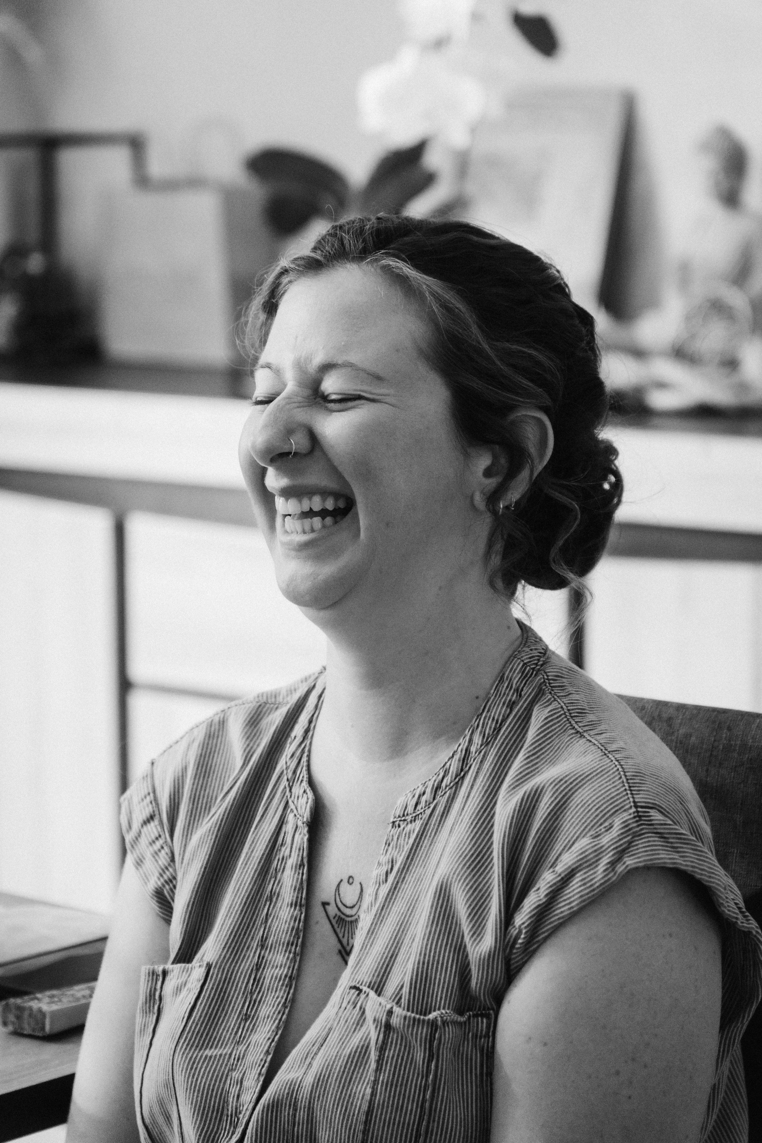 Une femme souriante avec un tatouage visible sur le haut de sa poitrine, assise à une table dans un intérieur, en noir et blanc.