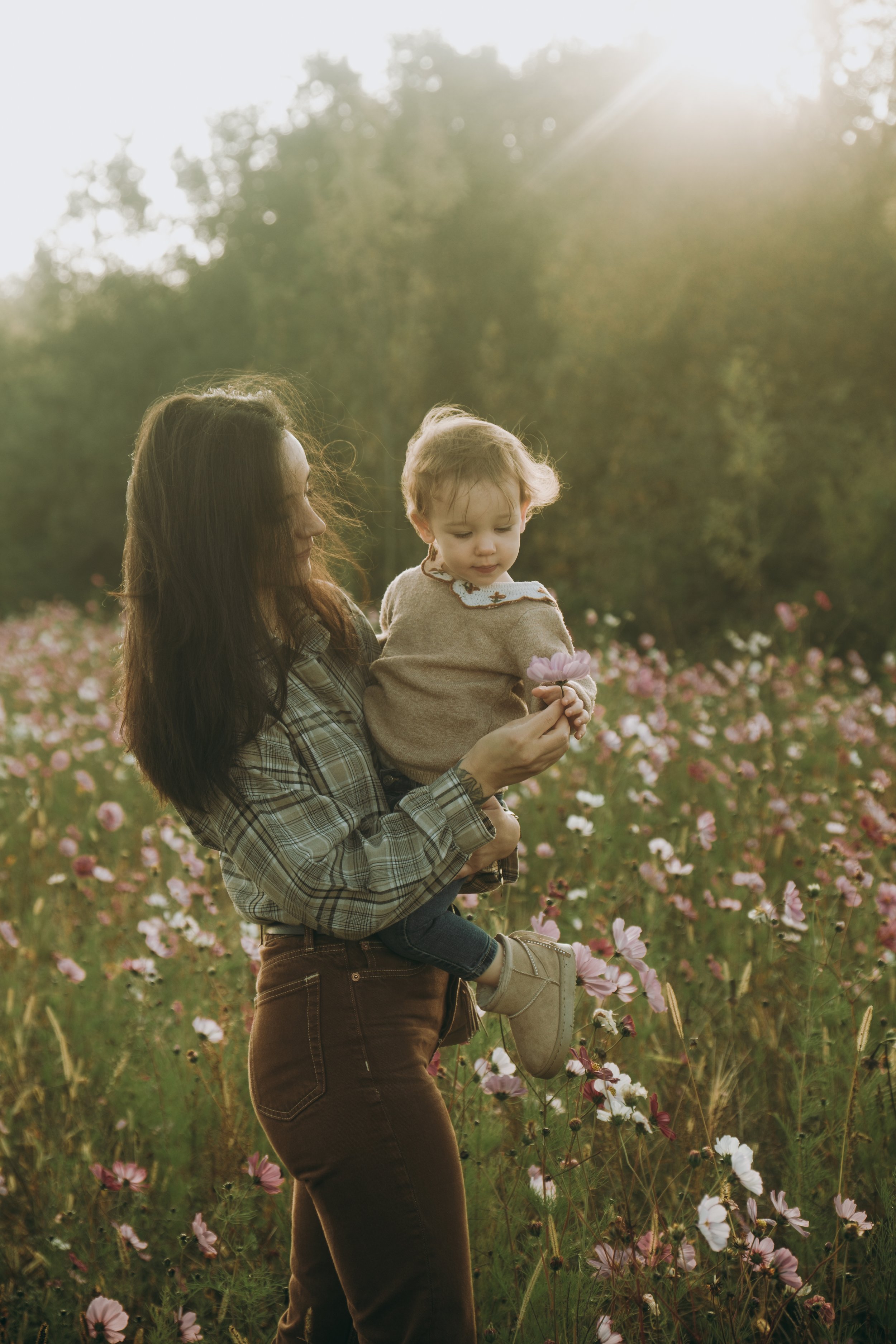Une femme tenant un enfant devant un champ de fleurs avec la lumière du soleil brillantes en arrière-plan.