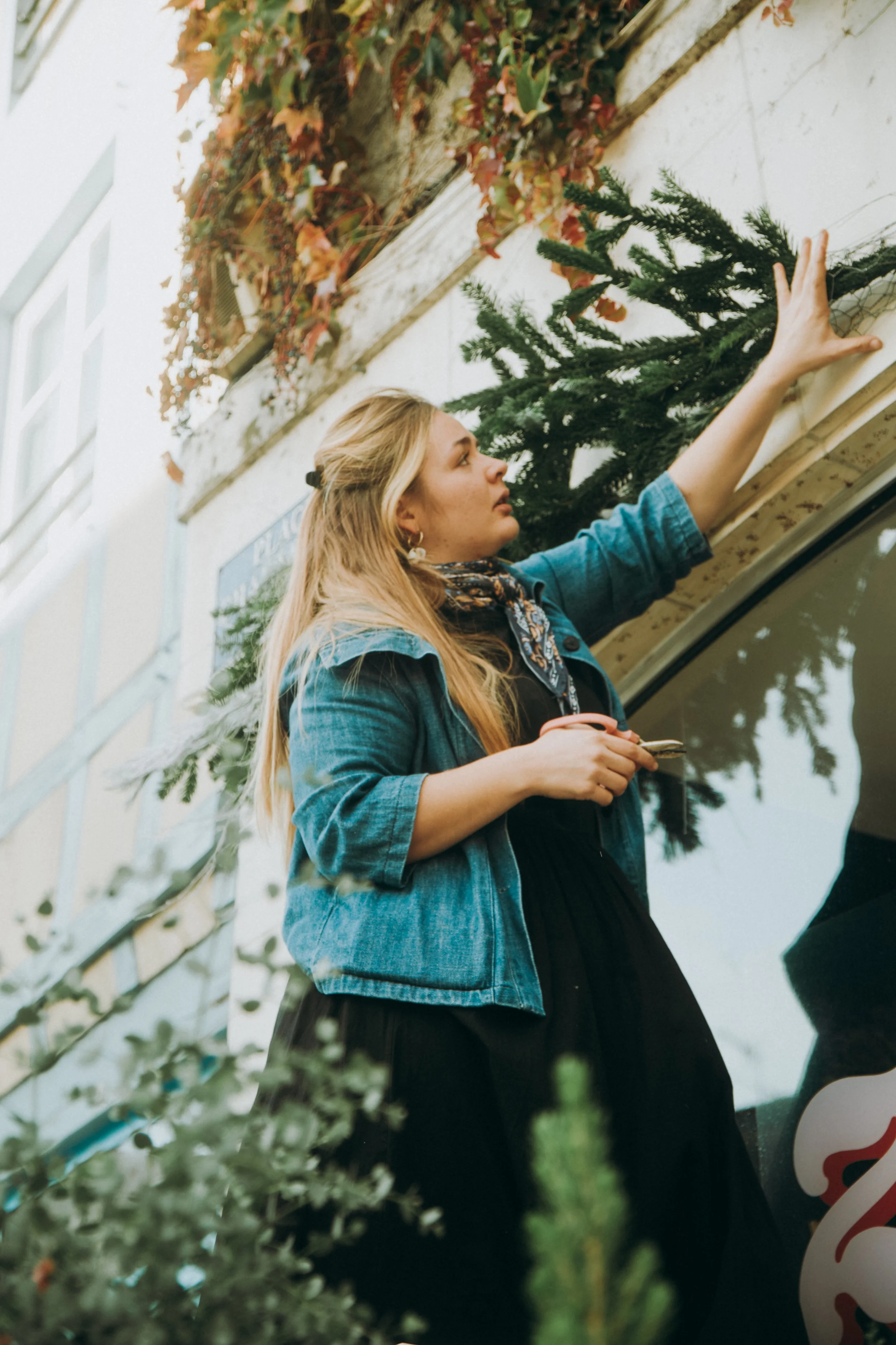 Une jeune femme avec des cheveux blonds, portant une veste en jean et un foulard, touche une plaque végétale sur un mur, avec des branches de sapin derrière elle et un véhicule en premier plan.