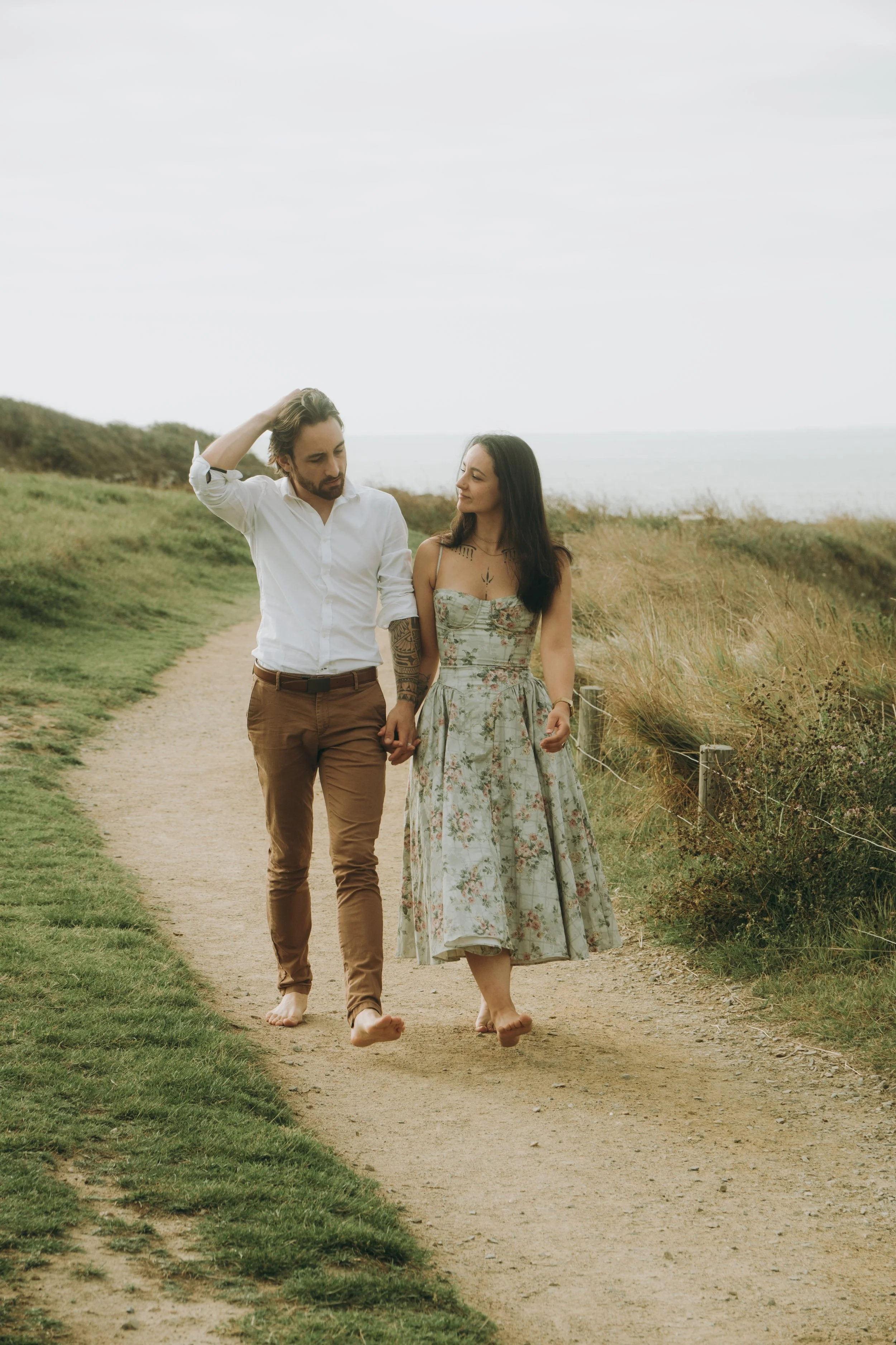 Un couple marche bras dessus bras dessous sur un sentier de campagne, tous deux pieds nus, avec la mer en arrière-plan.
