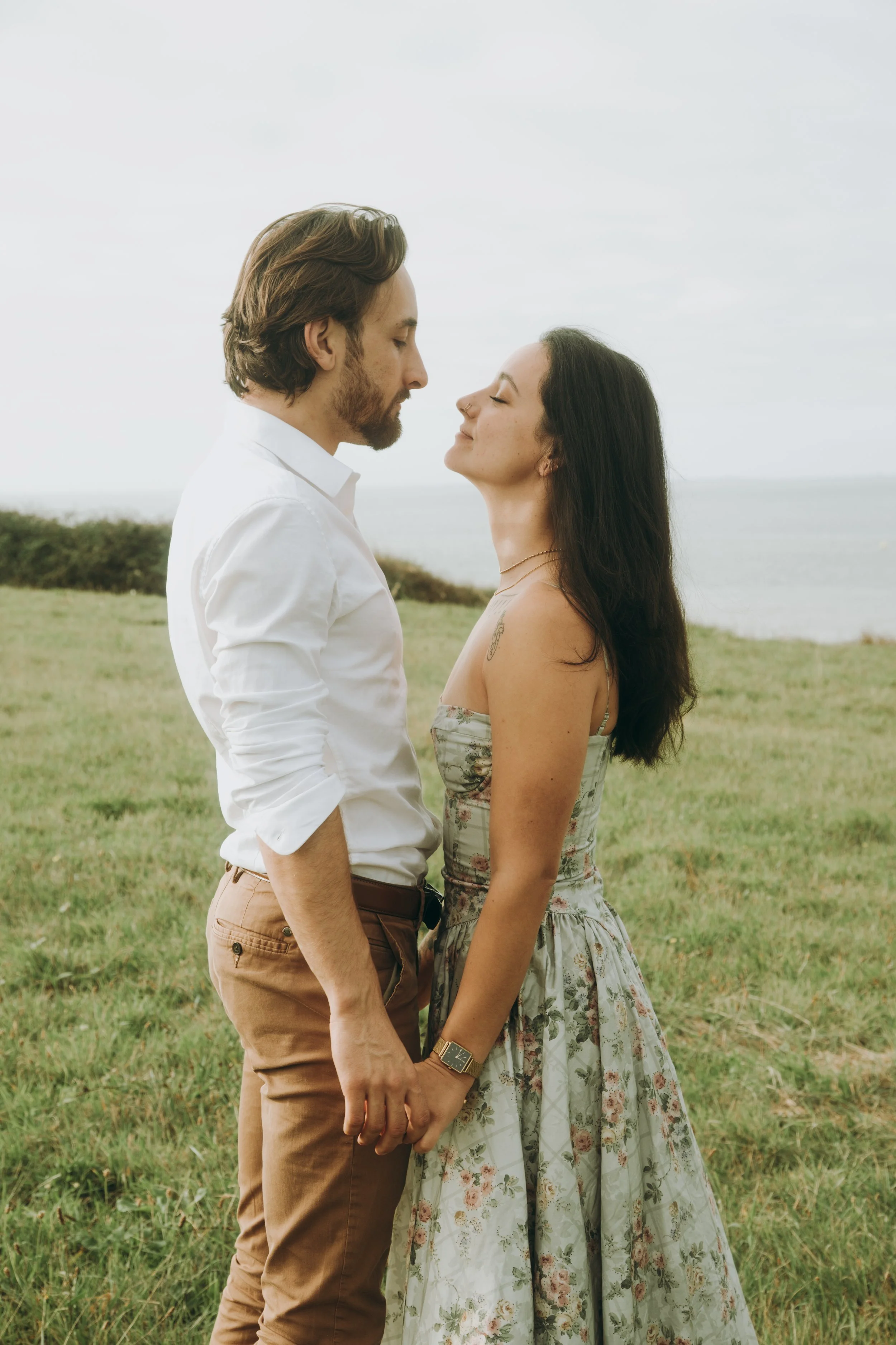 Un couple s'apprête à s'embrasser en tenant la main dans un espace vert près de la mer, avec un ciel nuageux en arrière-plan.