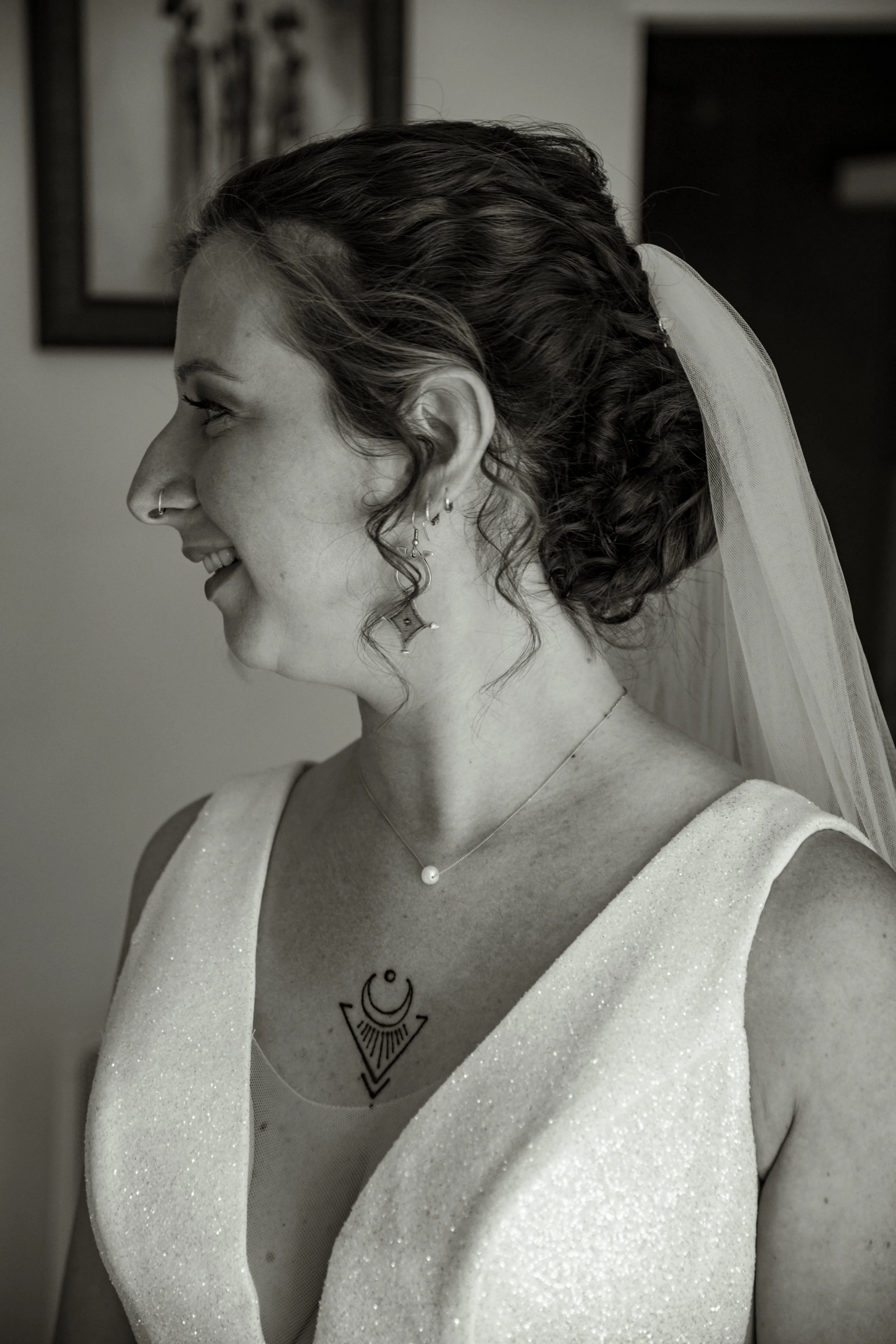 Une femme souriante avec des cheveux bouclés, portant un voile, des boucles d'oreilles, un collier avec une perle, et un tatouage graphique sur la poitrine, en profil, en intérieur.