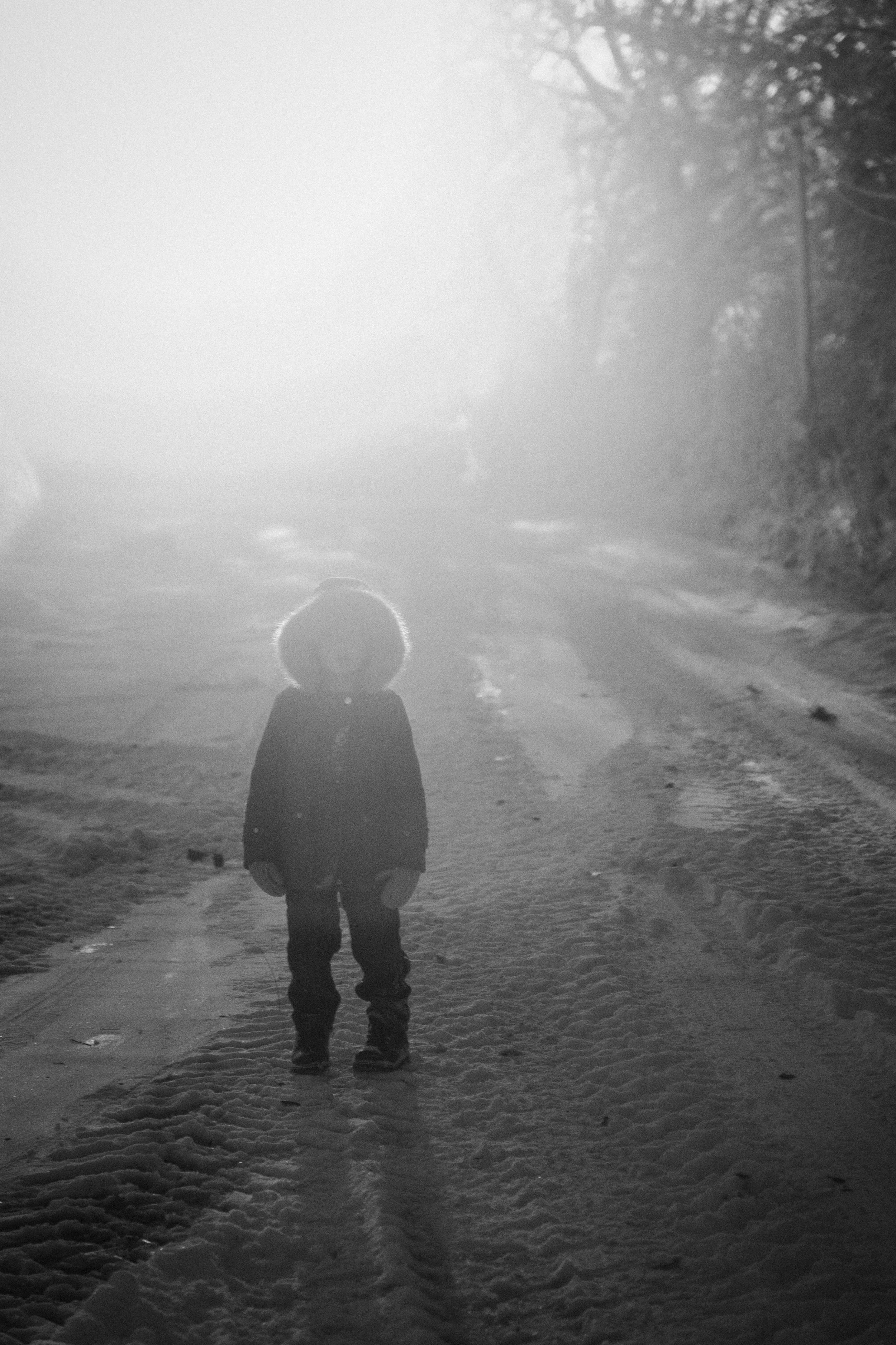 Un enfant portant un manteau à capuche dans une rue enneigée, avec une lumière forte à l'arrière-plan. La scène est en noir et blanc.