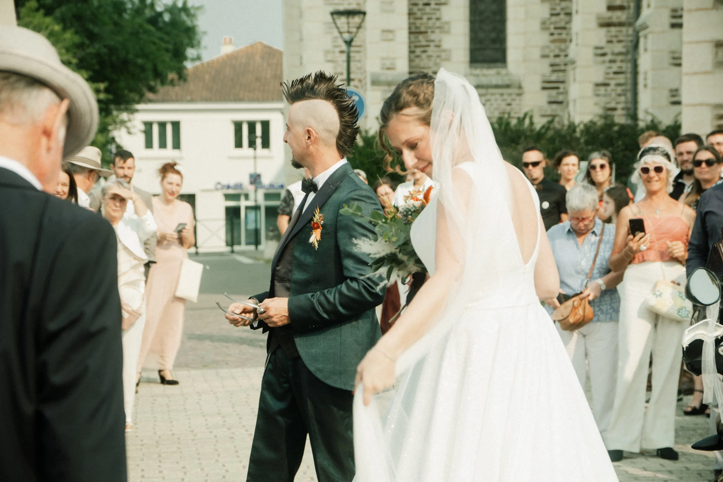 Un couple de mariés lors de leur mariage, entourés de spectateurs en plein air, avec une église en arrière-plan.
