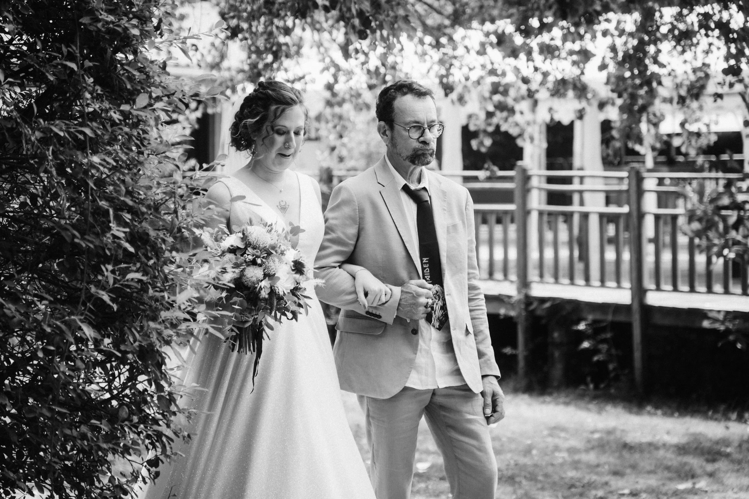 Une femme en robe de mariée tenant un bouquet, accompagnée par un homme en costume, lors d'une cérémonie ou d'une sortie dans un cadre extérieur.