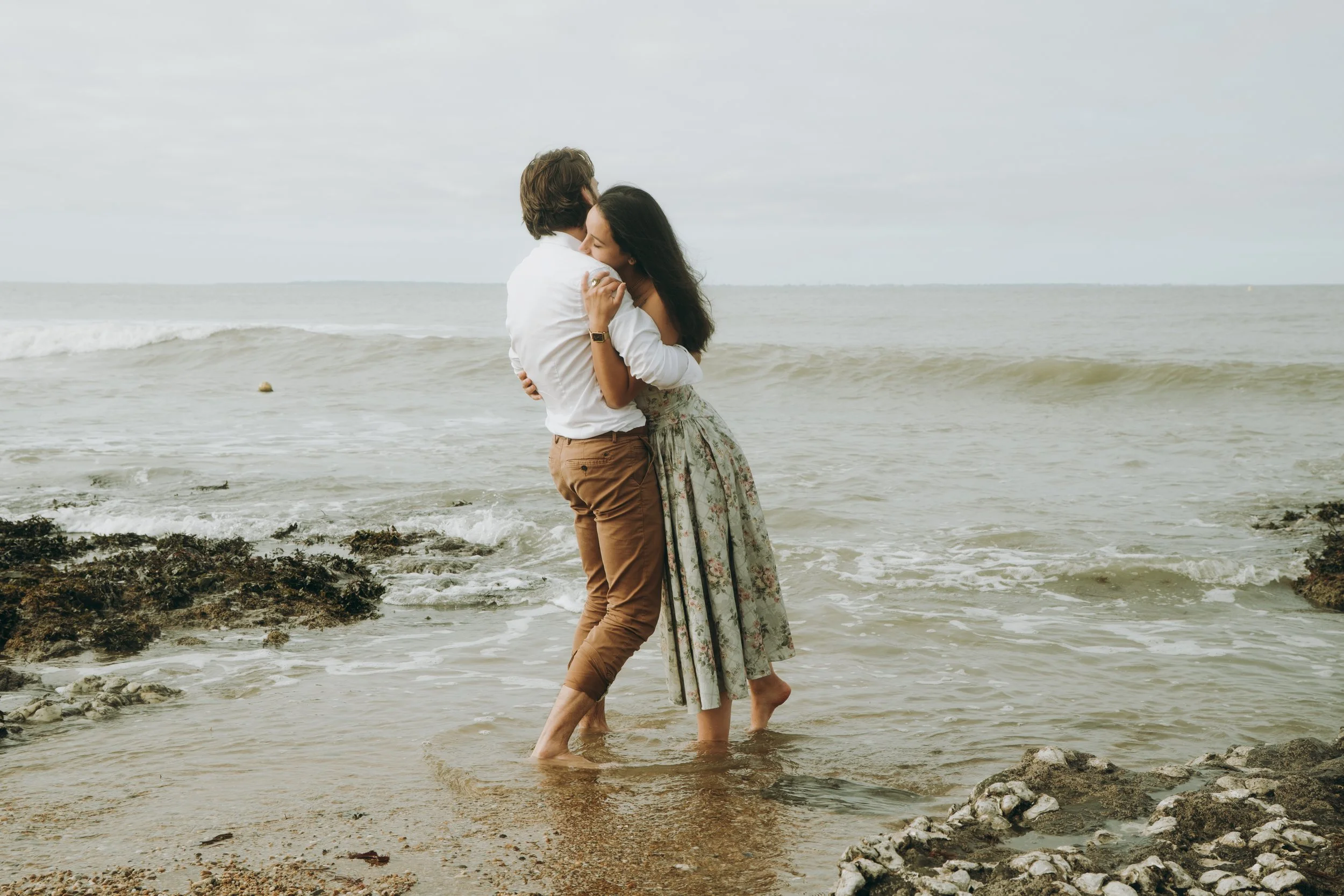 Un couple embrassé et se tenant par la taille, debout dans la mer peu profonde, à la plage, avec des rochers et des vagues en arrière-plan.