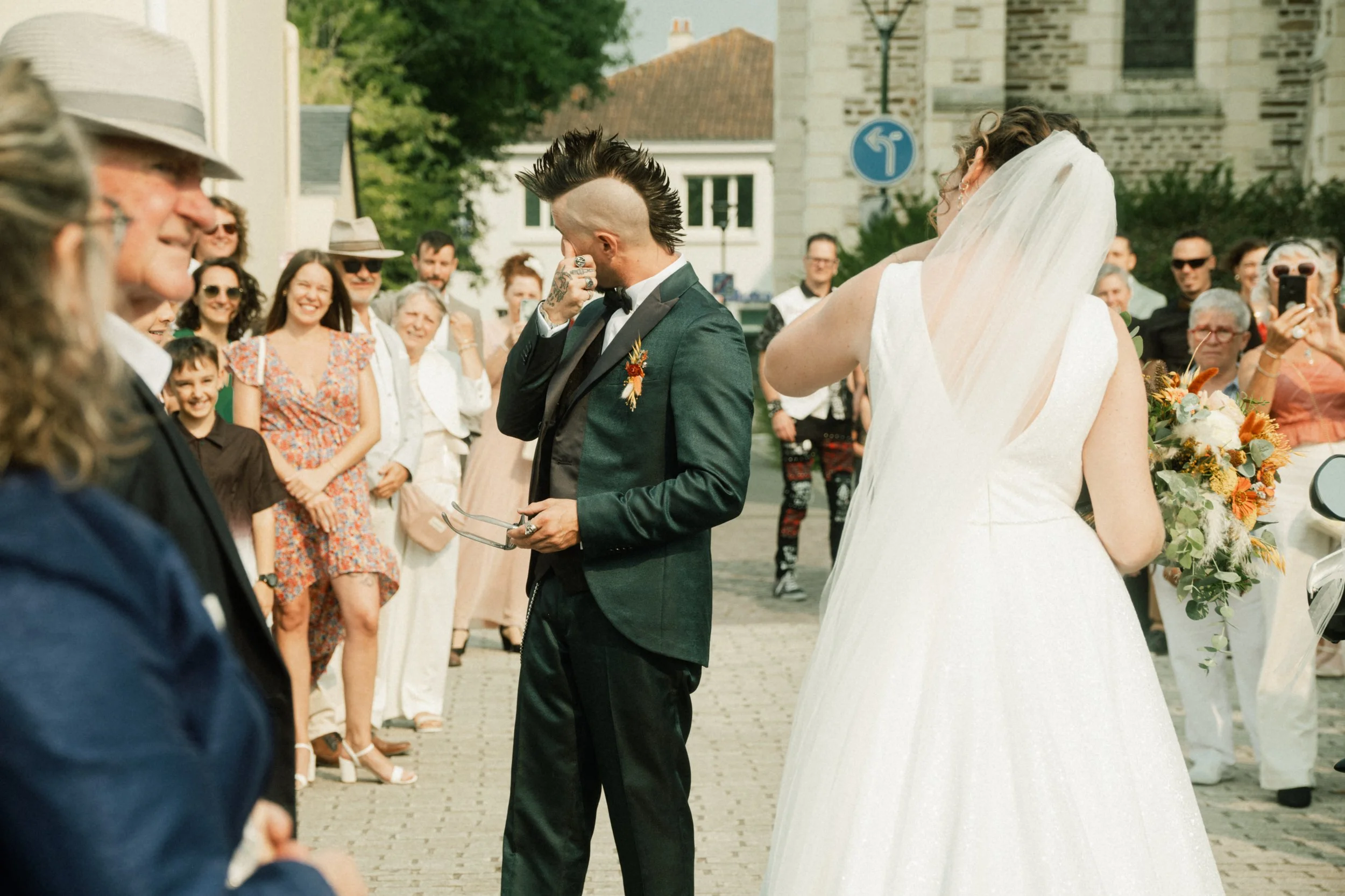 Un mariage en plein air avec un homme en costume noir et une femme en robe blanche, entourés de futurs mariés et de invités qui sourient.
