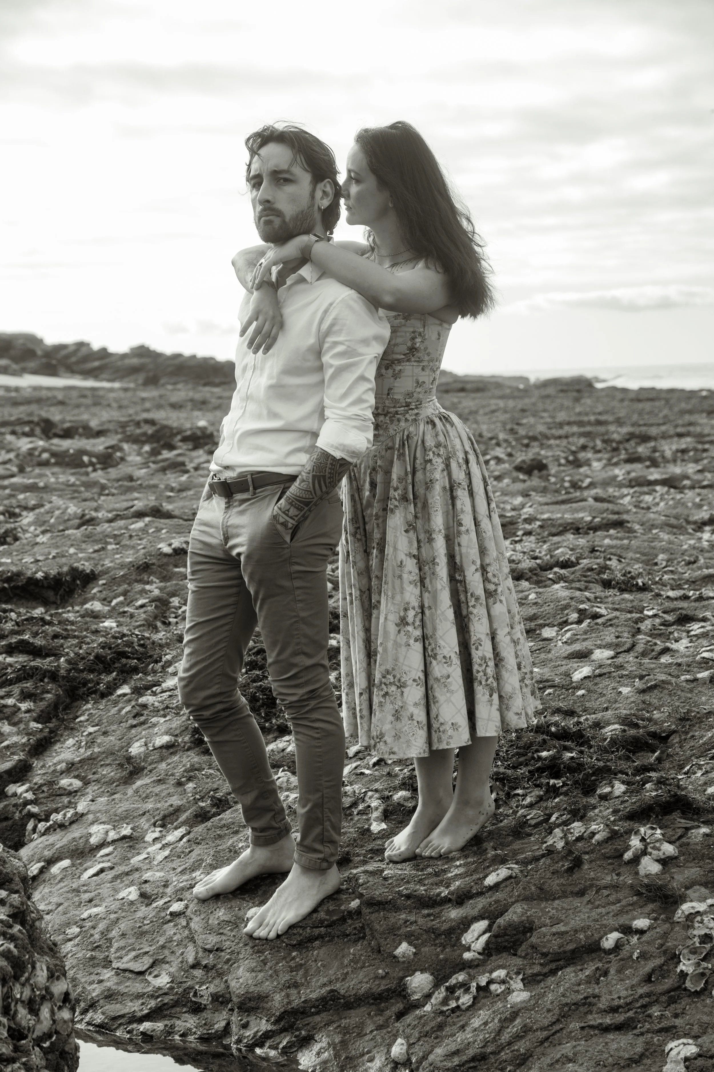 Un homme et une femme debout sur des rochers nus et plats au bord de la mer. La femme embrasse l'homme sur la poitrine. La photo est en noir et blanc, le ciel est partiellement nuageux.