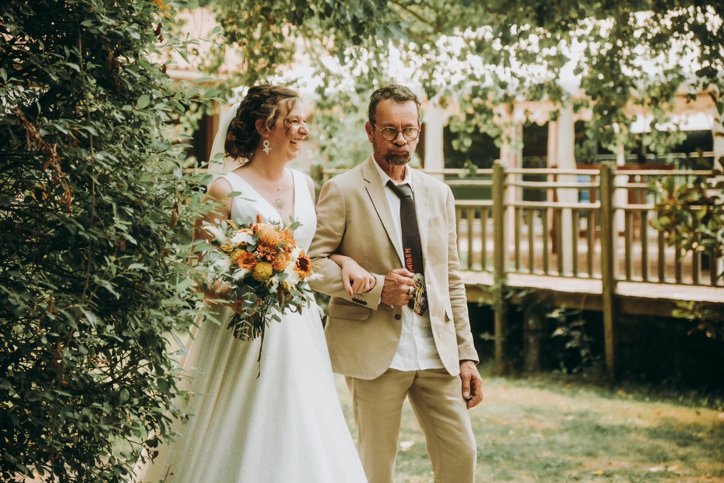 Un couple de mariés marche dans un jardin, la mariée porte une robe blanche et tient un bouquet de fleurs. Elle sourit tandis que l'homme, en costume beige, marche à côté d'elle.