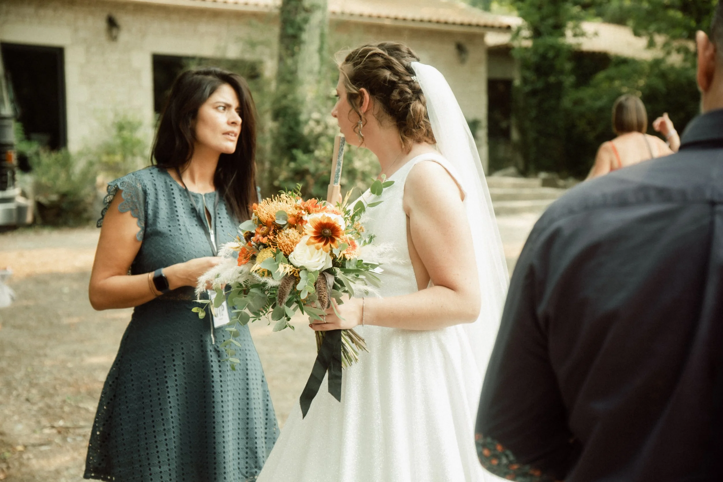 Deux femmes en discussion lors d'un mariage en plein air. La femme à gauche porte une robe bleue et la femme à droite porte une robe de mariée blanche, tenant un bouquet de fleurs.