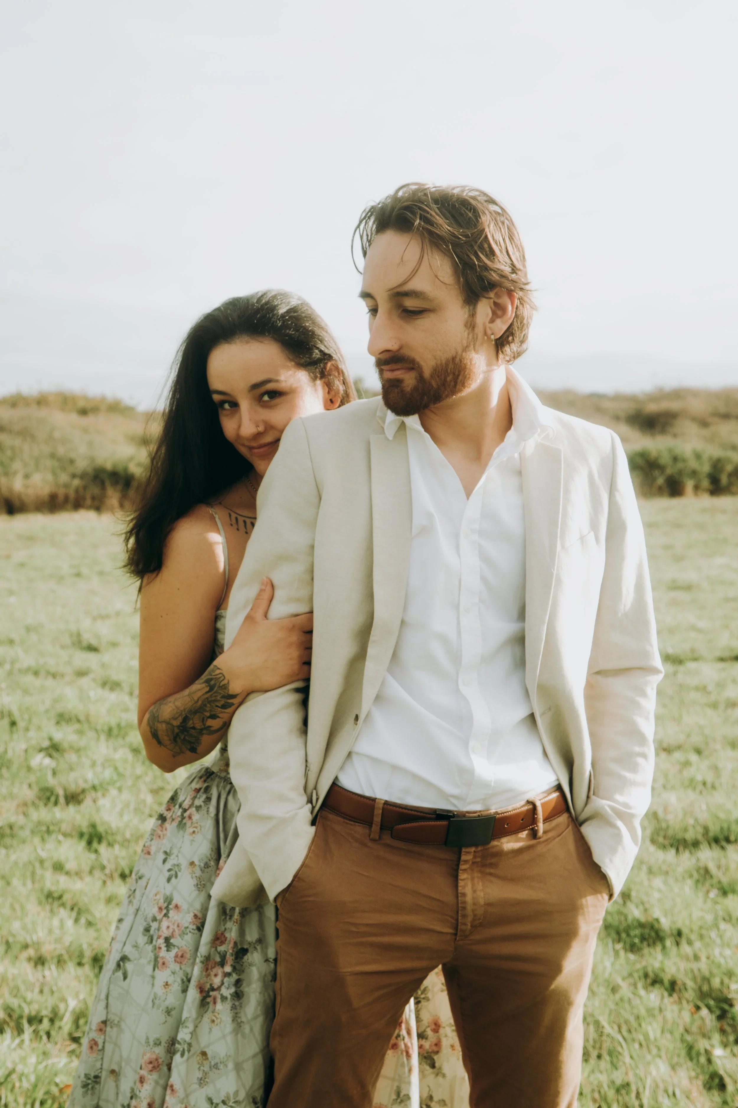 Un homme et une femme posant ensemble dans un champ, l'homme porte un costume beige et des pantalons marron, la femme porte une robe fleurie, tous deux semblent détendus et souriants.