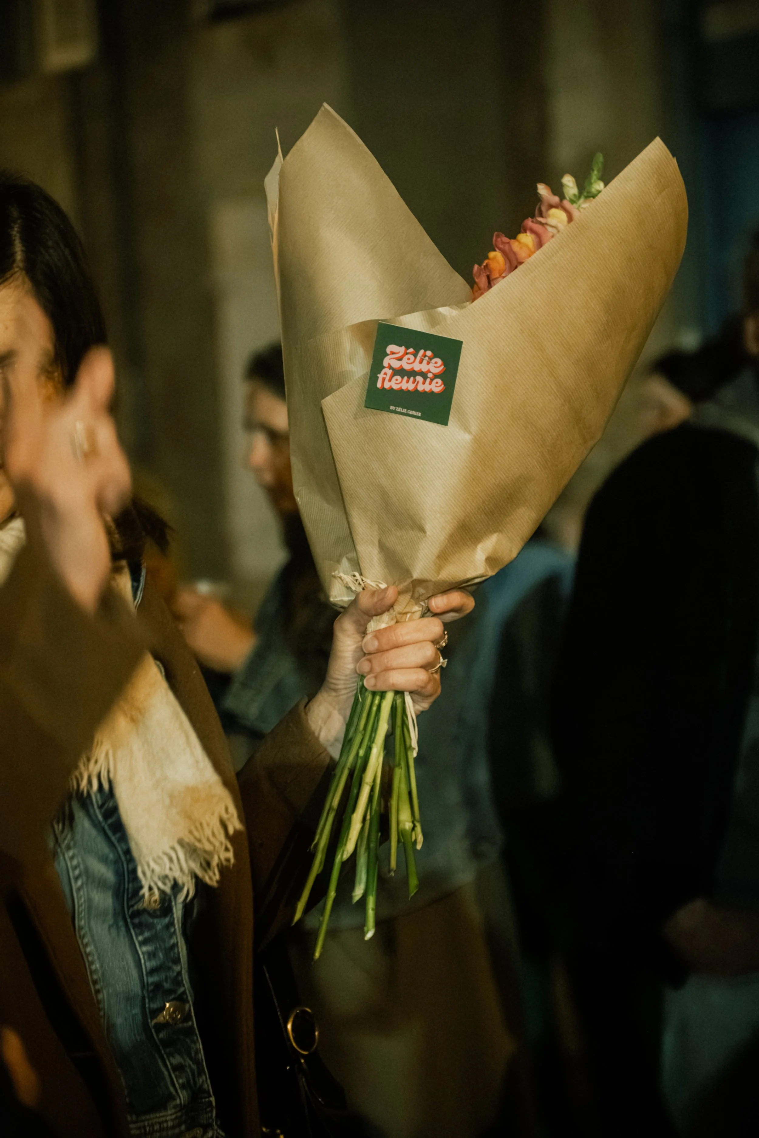 Une personne tient un bouquet de fleurs enveloppé dans du papier kraft. Le bouquet a une étiquette rouge et verte avec l'inscription 'Zélie Fleurie'.