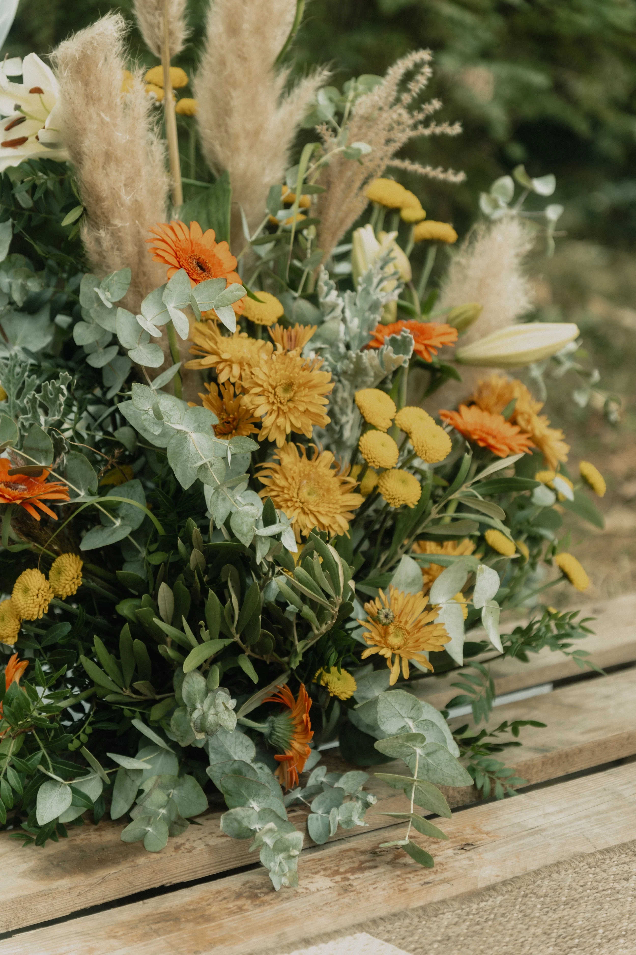 Un bouquet de fleurs comprenant des gerberas jaunes et oranges, des lys blancs, de la lavande, des feuilles d'eucalyptus et des herbes pampas, posé sur une surface en bois.