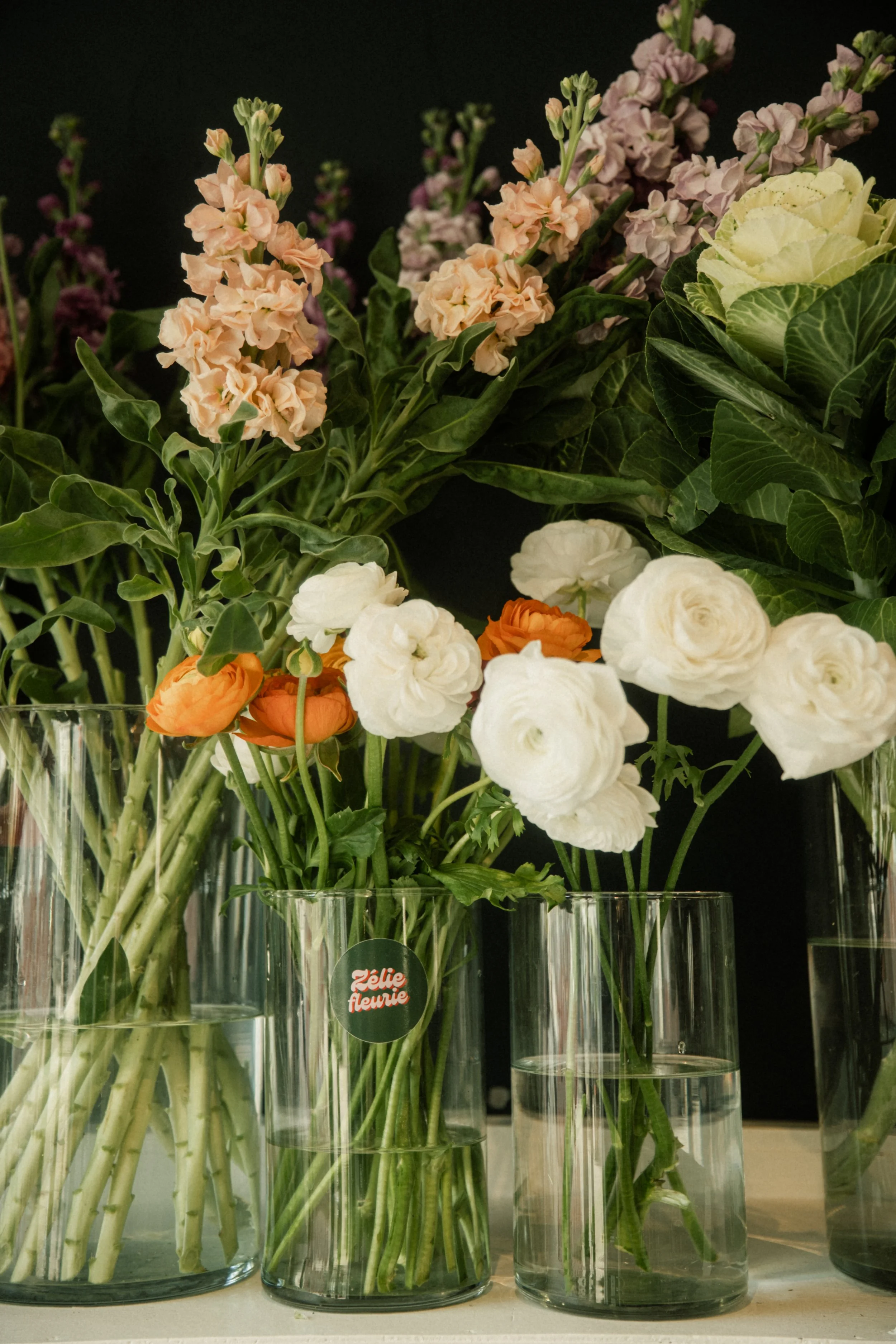 Fleurs dans des vases en verre, incluant des œillets blancs, des renoncules orange et des fleurs violettes et roses, sur une table.