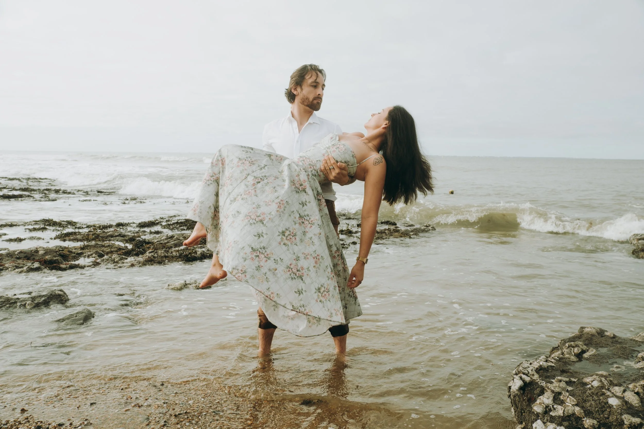 Un homme en chemise blanche tenant une femme habillée en robe longue à fleurs au bord de la mer, l'eau jusqu'aux jambes. Le ciel est nuageux.