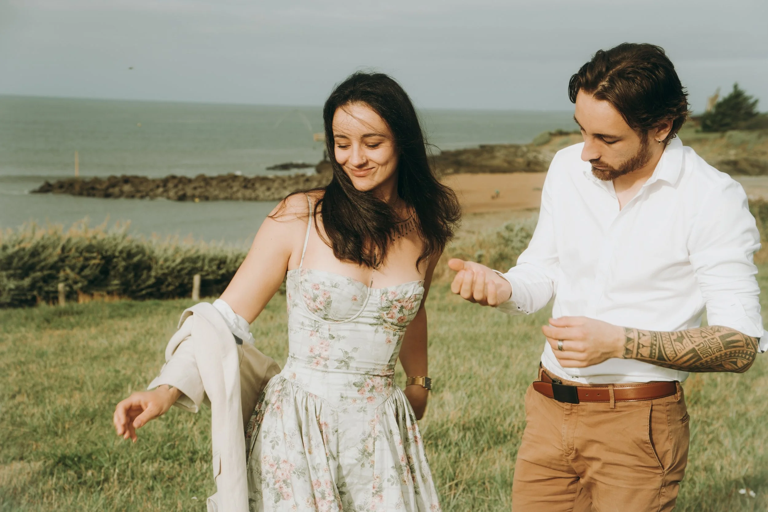 Un jeune homme et une jeune femme se promènent côte à côte près de la mer, dans un paysage marin avec de l'herbe et des rochers. La femme porte une robe à fleurs et sourit tandis que l'homme, en chemise blanche et pantalon beige, semble discuter avec