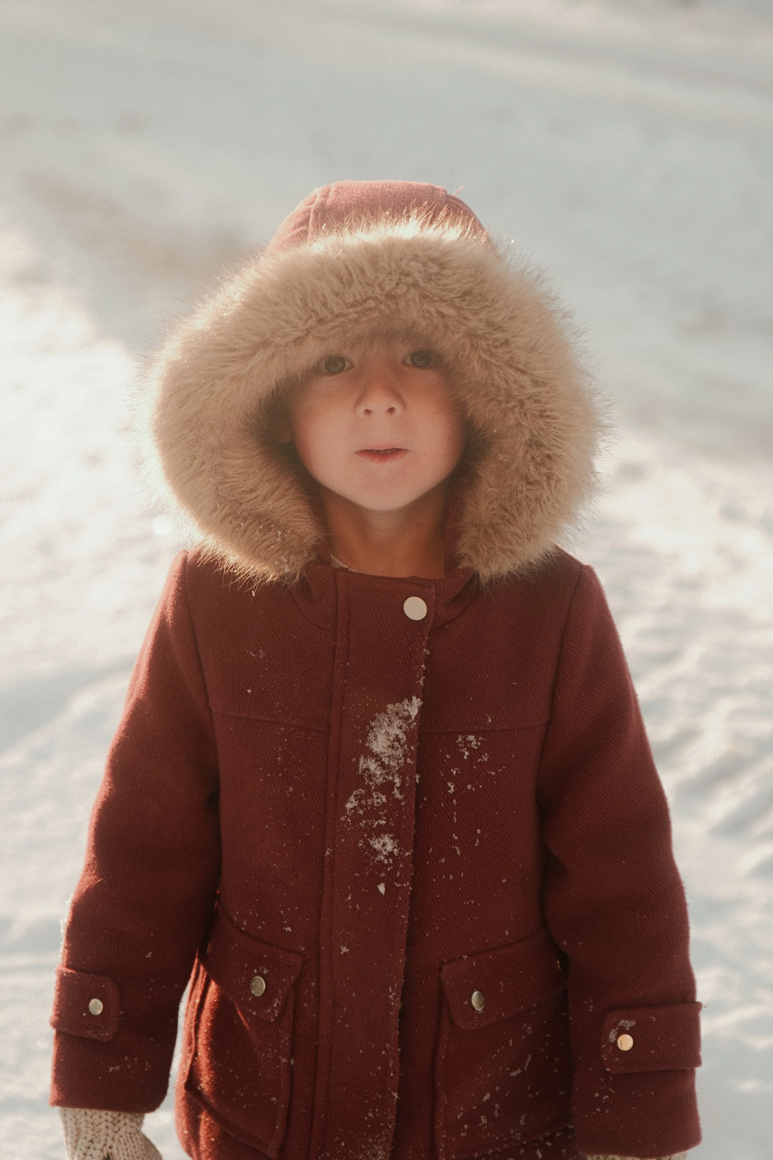 Un jeune enfant vêtu d'un manteau rouge avec une capuche en fourrure, debout dans la neige.