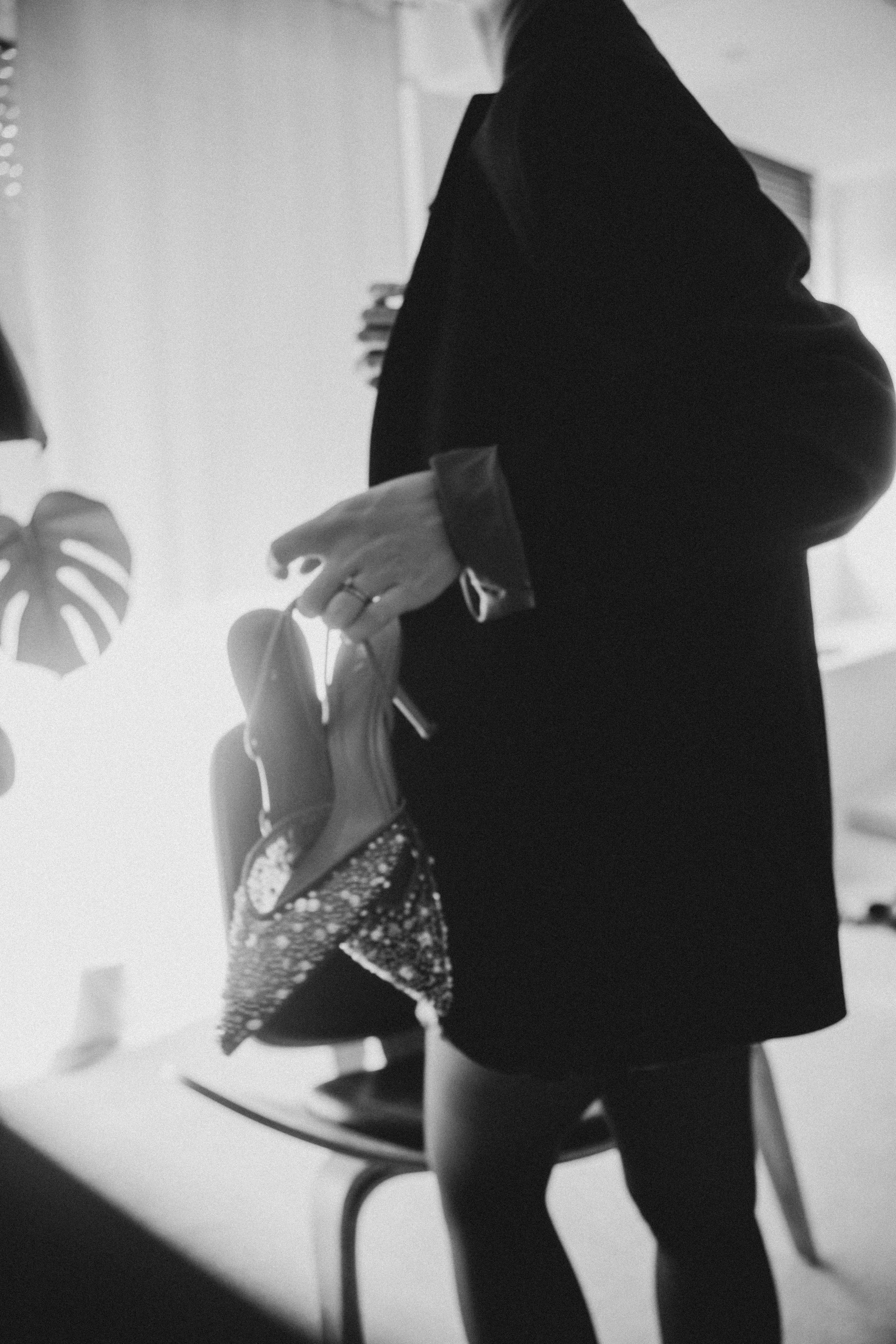 Une femme en manteau noir et pantalon tient un sac à main, dans un intérieur moderne avec des plantes dans le fond.