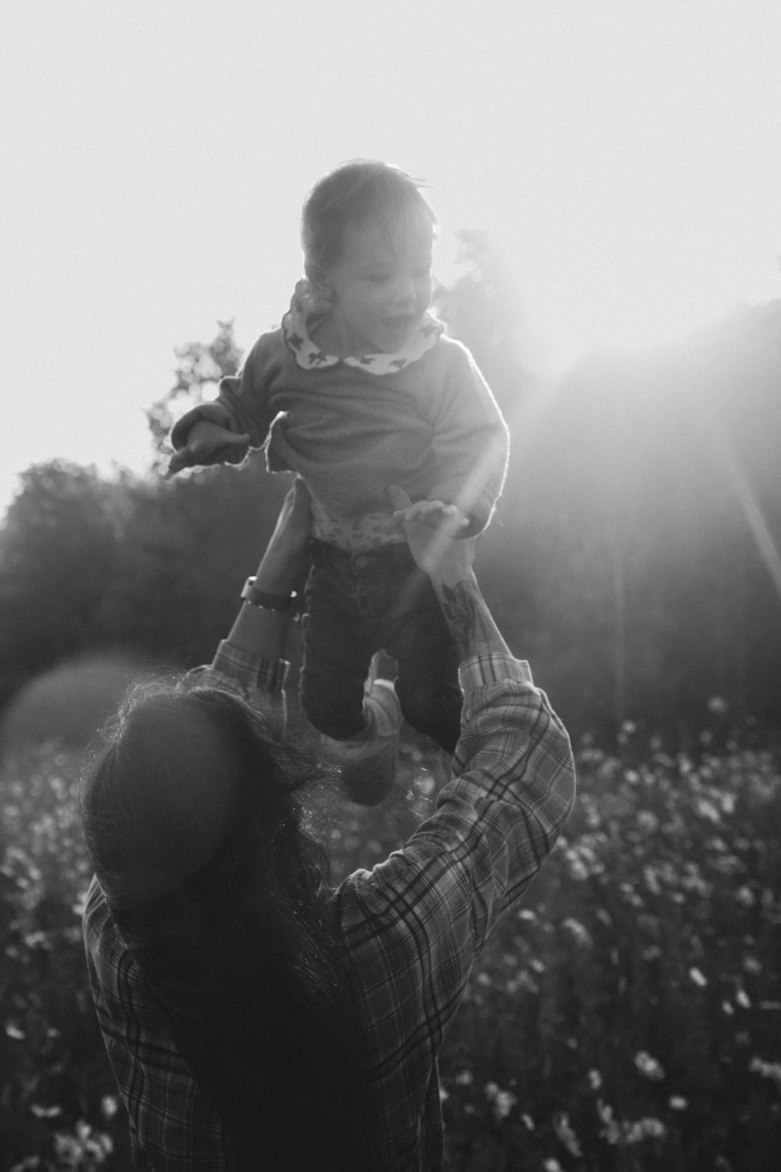 Une personne qui lève un enfant en l'air lors d'une sortie en plein air, avec le soleil brillant derrière eux.