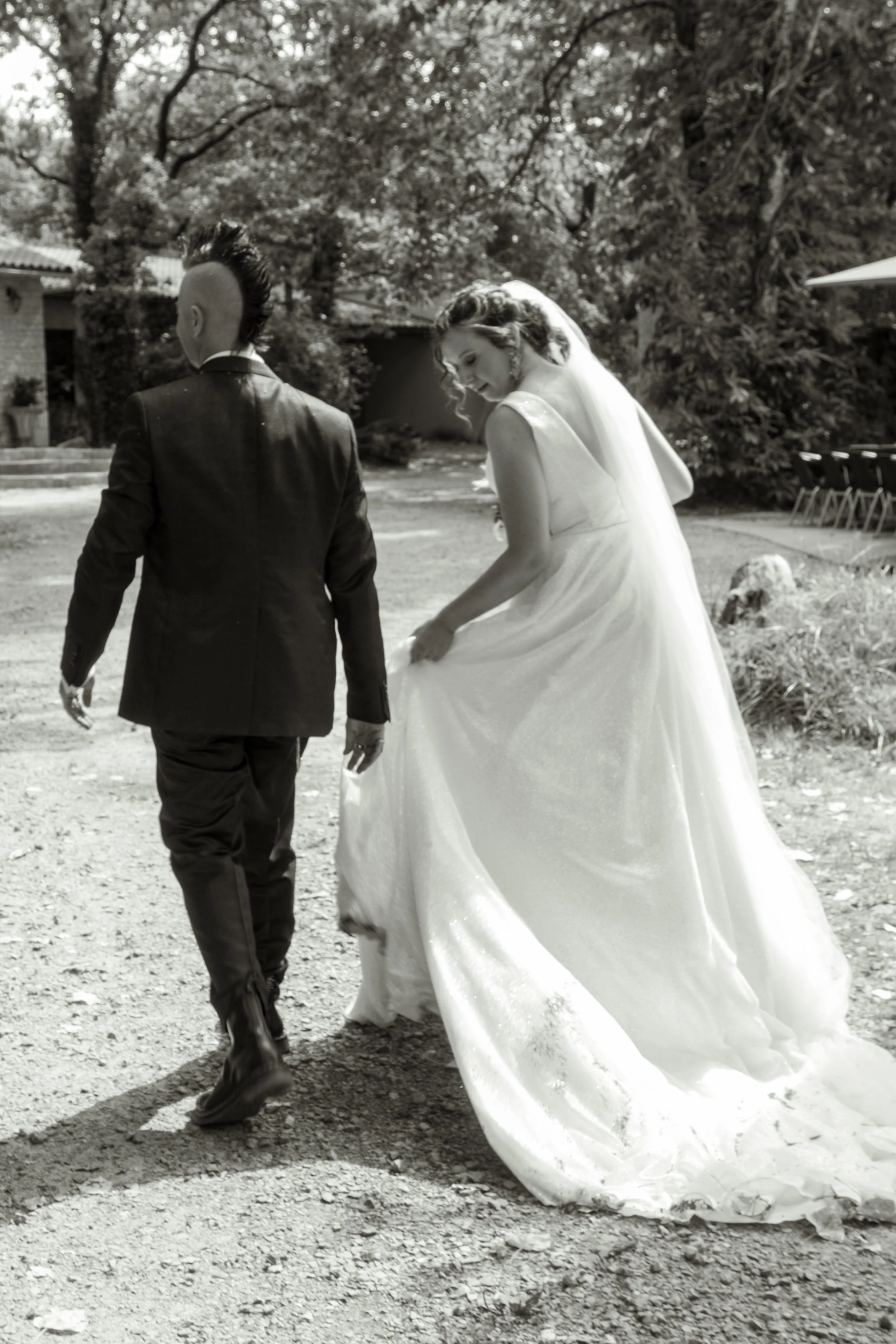 Un couple de mariés marche dans un jardin, la femme en robe blanche et le homme en costume noir, en noir et blanc.