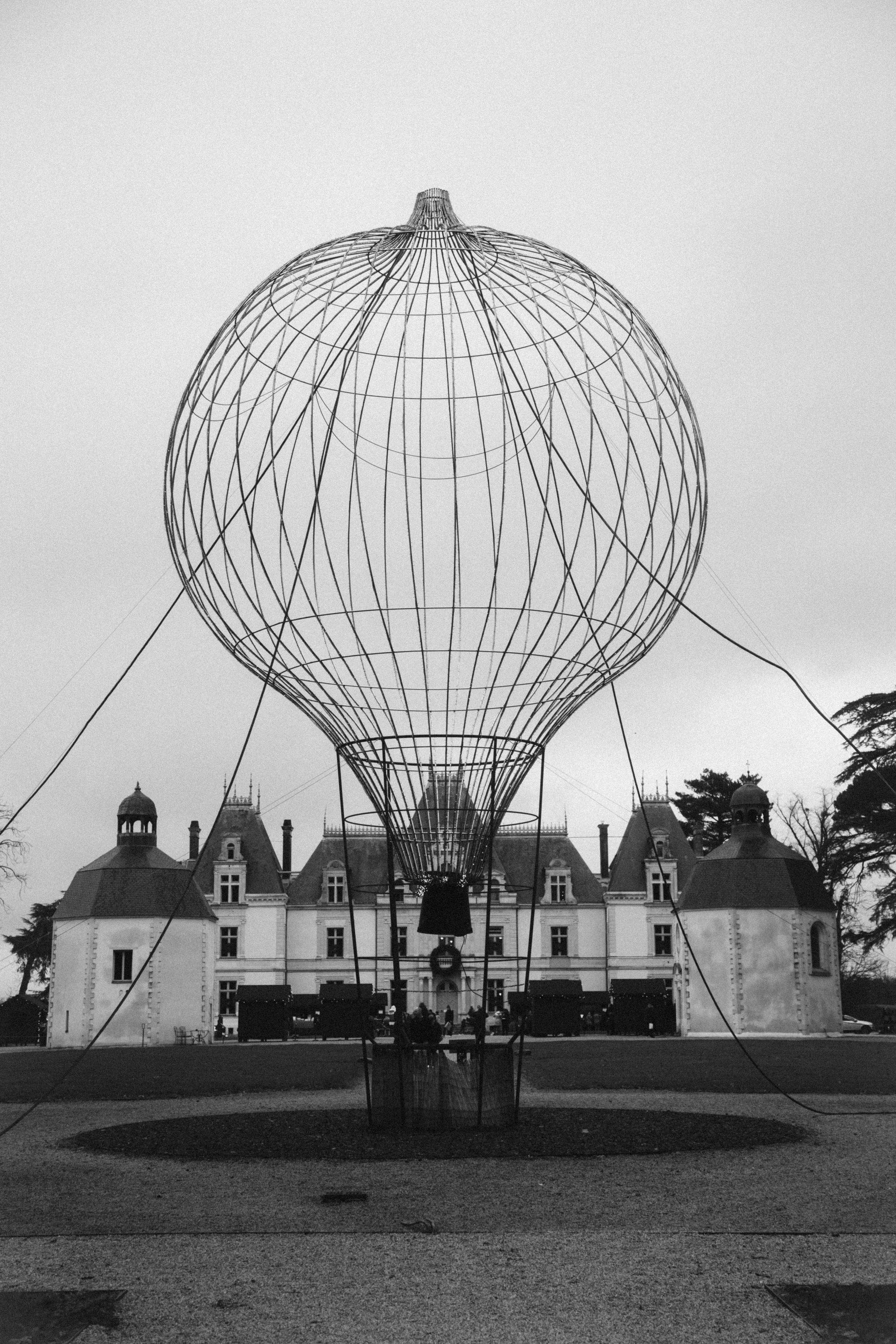 Une structure de montgolfière en fil métallique en forme de ballon dans un parc devant un château à l'arrière-plan, en noir et blanc.