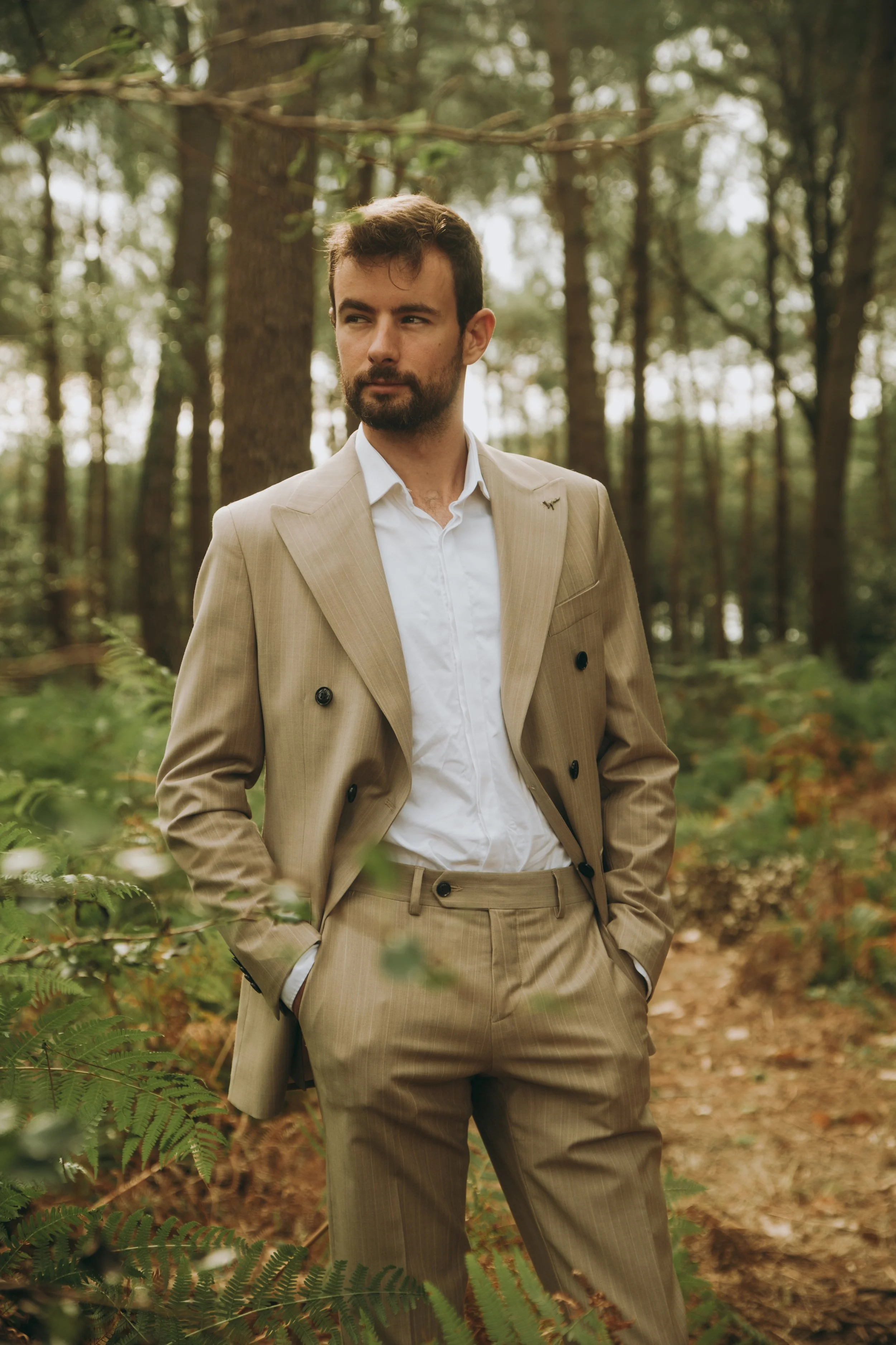 Un homme habillé en costume beige, debout dans une forêt, en regardant sur le côté.