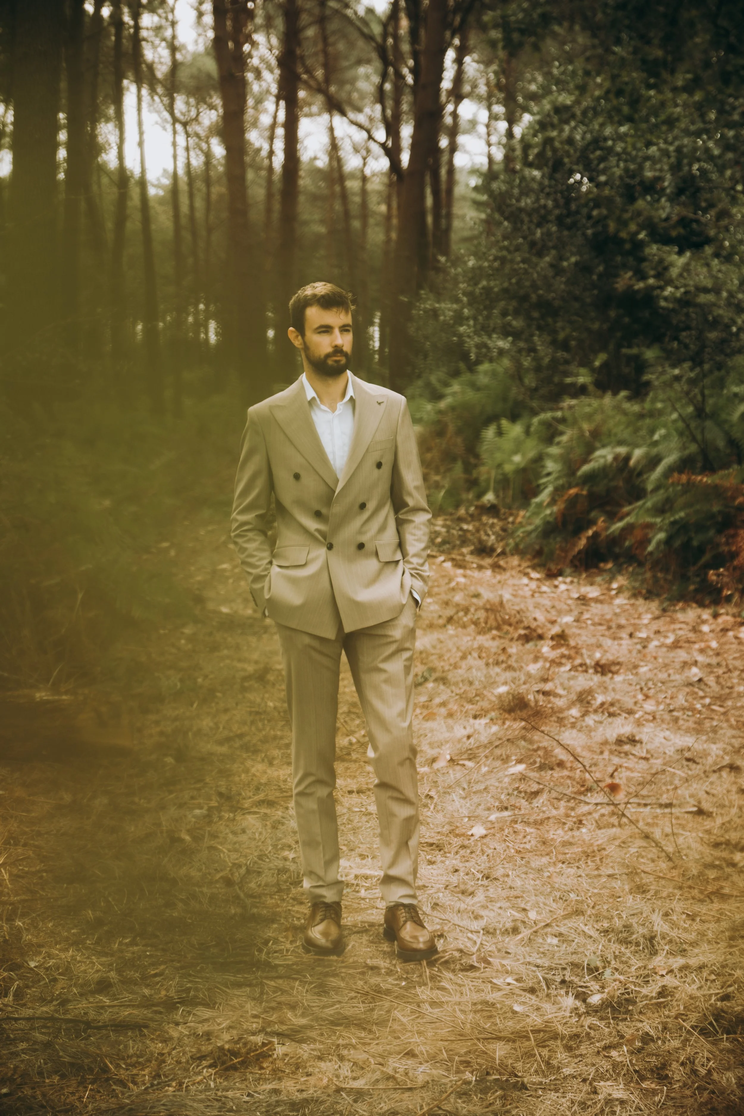 Un homme en costume beige marche dans une forêt, mains dans les poches, regardant vers la gauche.