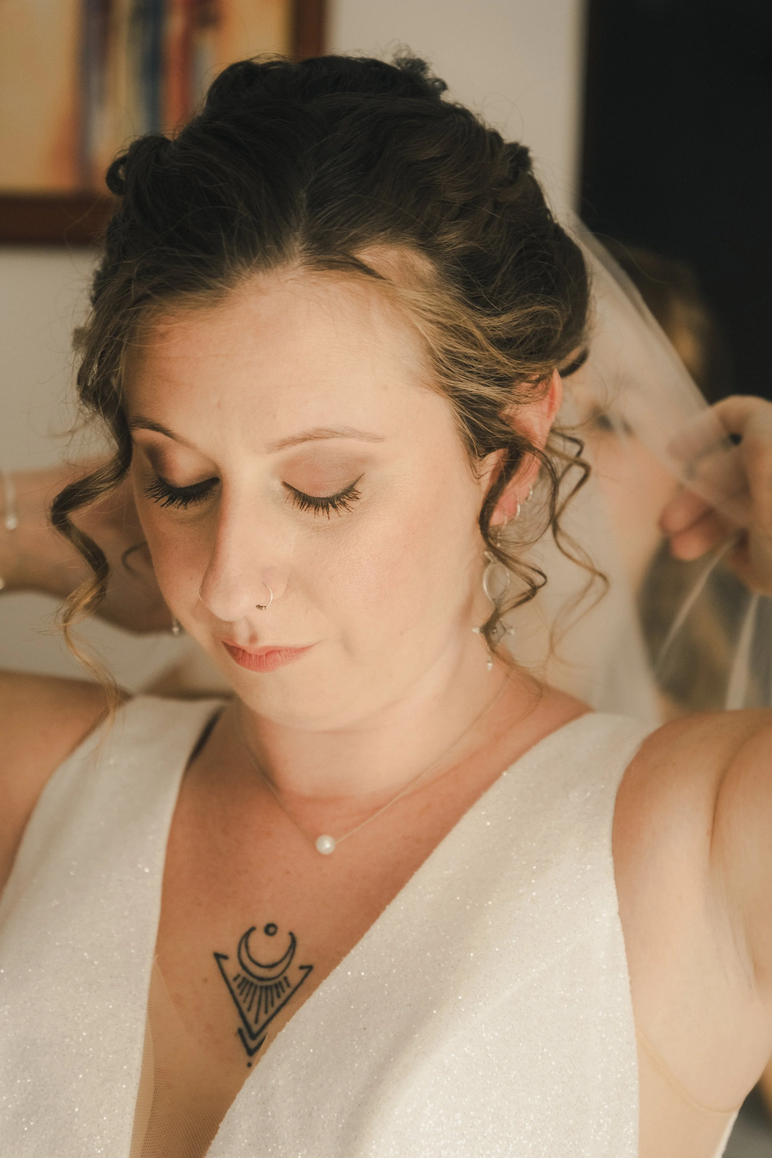 Une femme, probablement une mariée, ajuste sa coiffure ou son voile, avec des yeux fermés et un léger sourire. Elle porte une robe blanche, des bijoux en perles et un tatouage noir dans le décolleté.