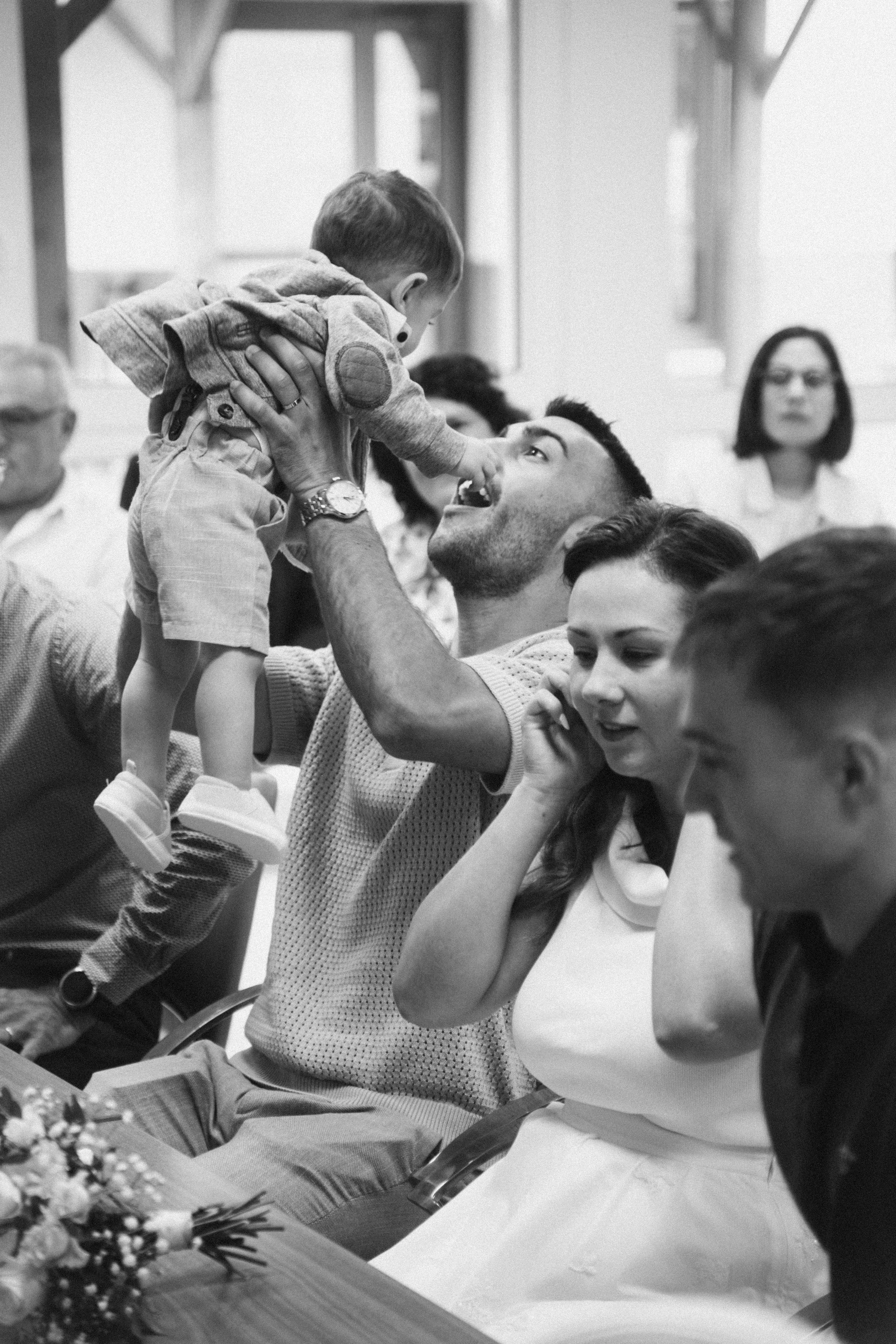 Un homme tient un jeune garçon élevé en l'air lors d'une réunion ou célébration, avec d'autres personnes assises autour.
