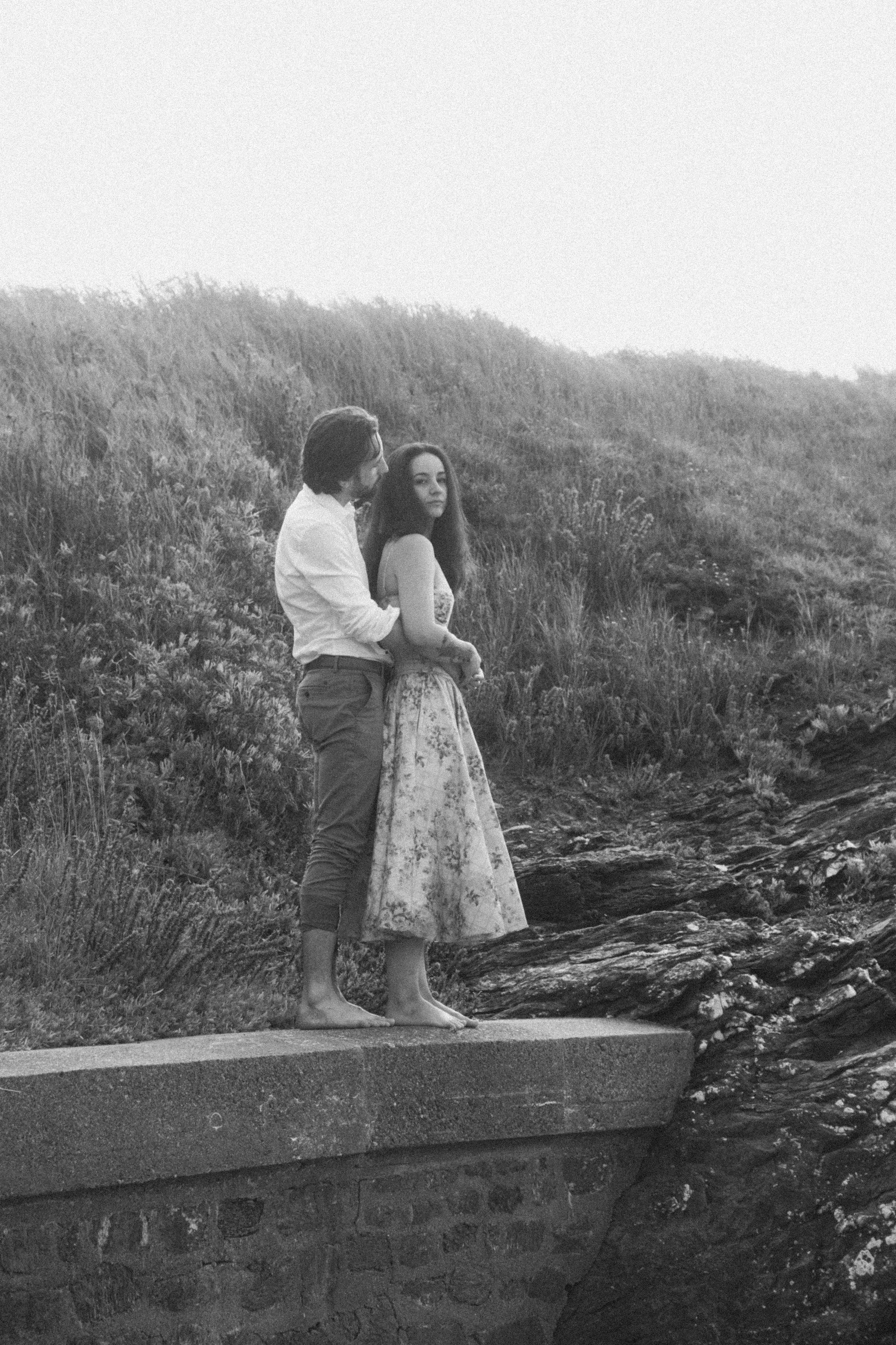 Un couple debout sur un muret en pierre, près de l'eau, dans un paysage naturel, vue en noir et blanc.