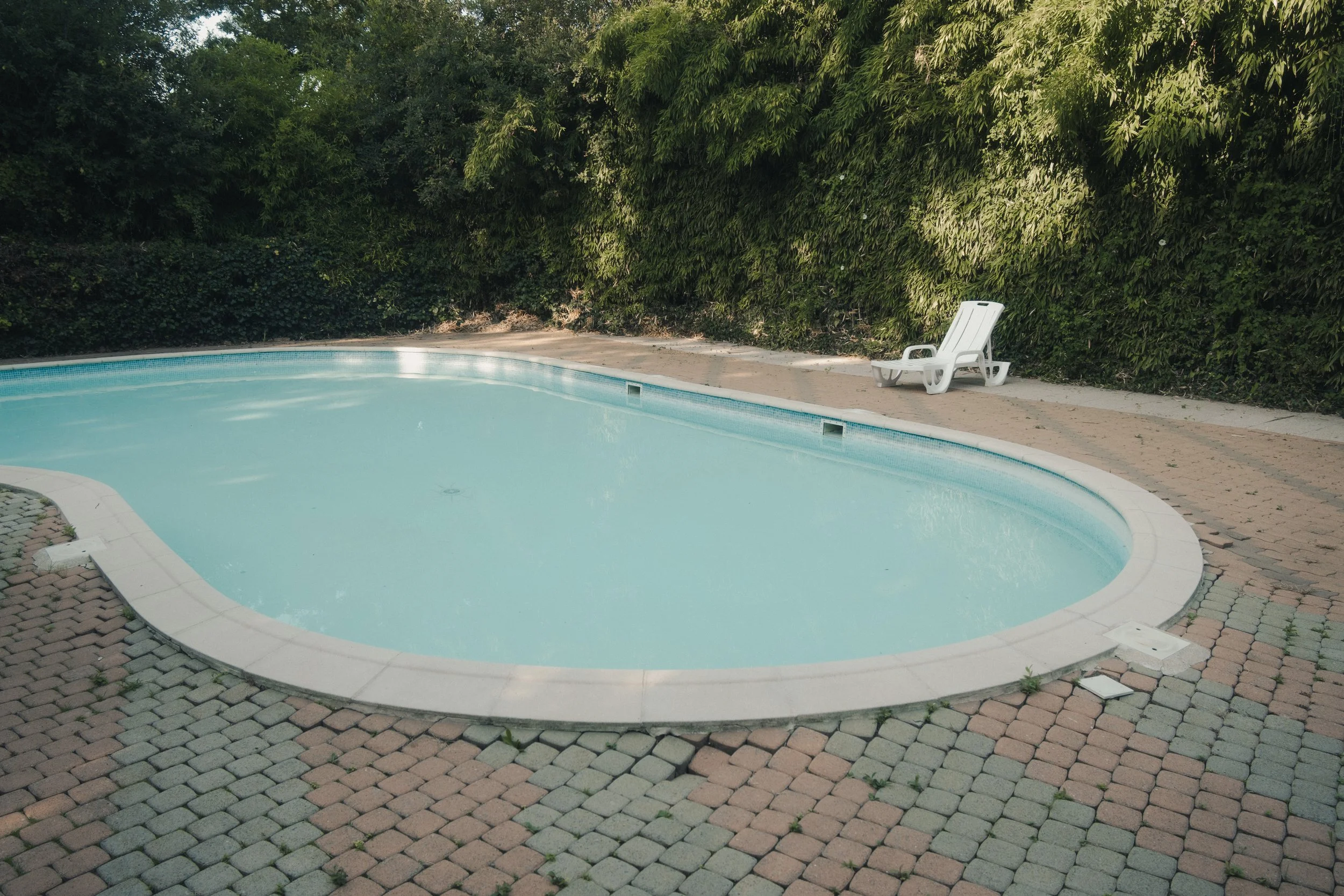 piscine vide et une chaise longue blanche sur une terrasse en pavés, avec un fond de buissons verts