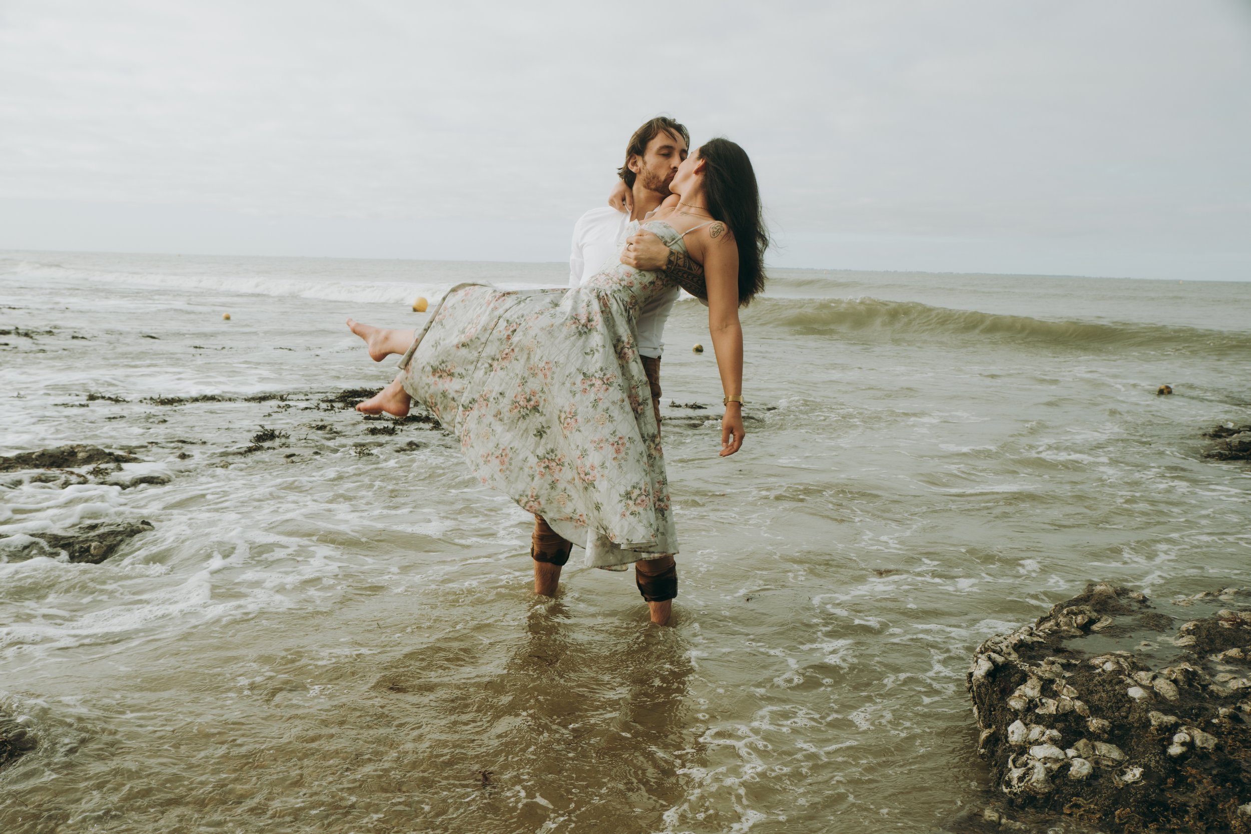 Un couple se tient dans l'eau de la mer, lui portant une femme dans ses bras. Elle porte une robe longue et lui un pantalon, tous deux pieds dans l'eau. La mer est calme avec quelques rochers visibles.