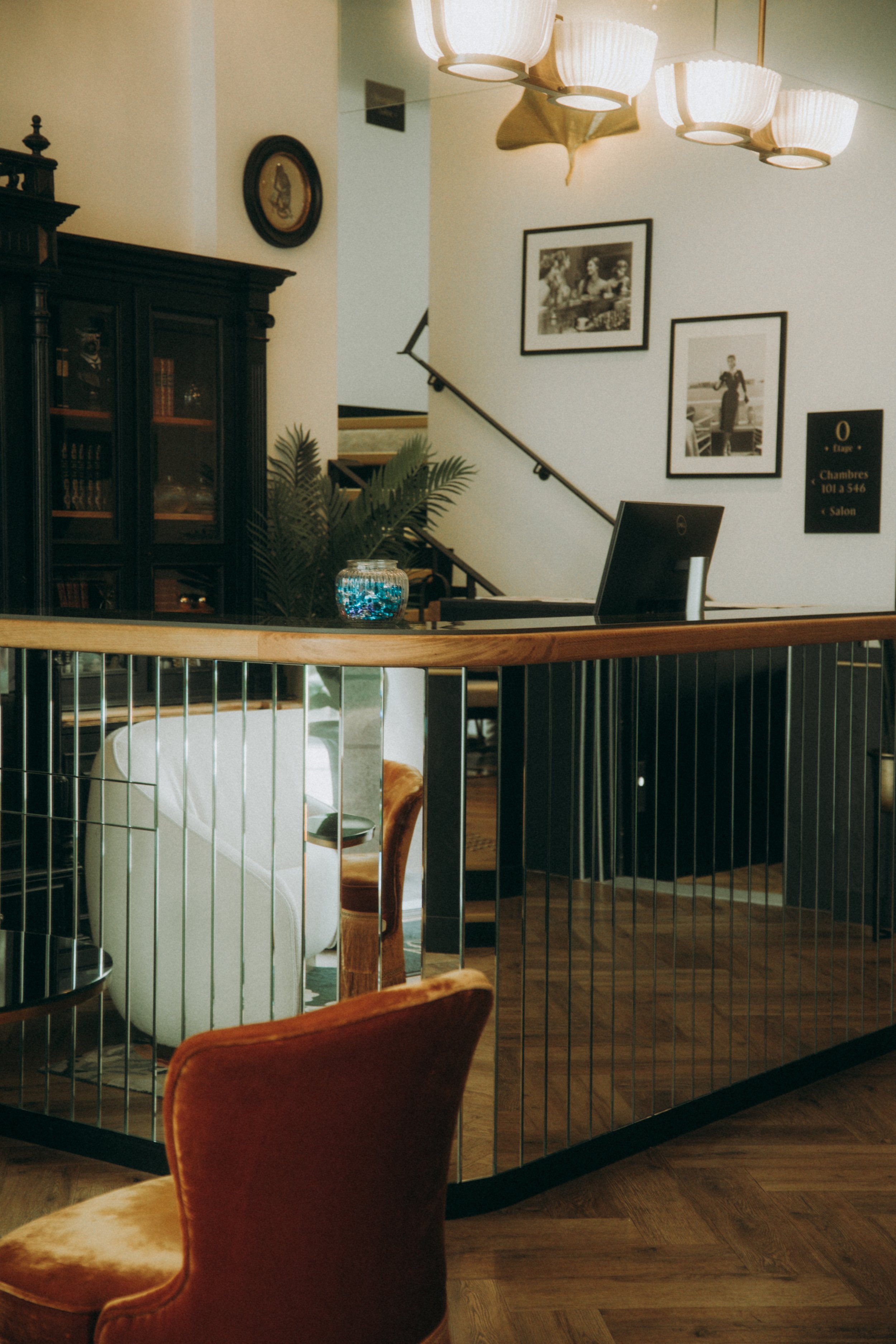 Vue de l'entrée d'un hôtel avec un comptoir, une chaise orange, une bibliothèque en bois sombre, des tableaux noirs et blancs, une lampe suspendue, et un escalier en arrière-plan.