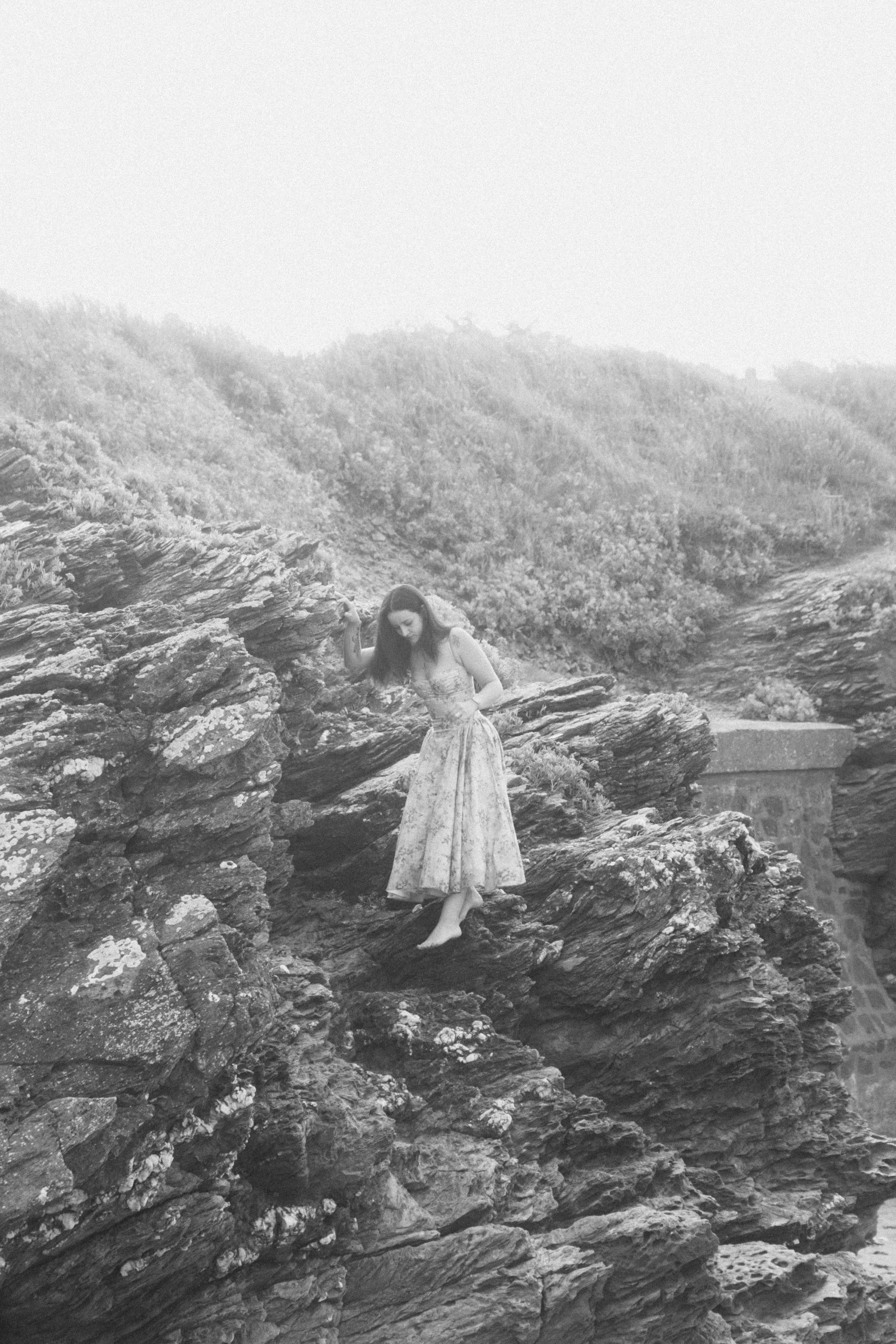 Une femme en robe longue se tient sur des rochers près de la mer, dans un cadre naturel.