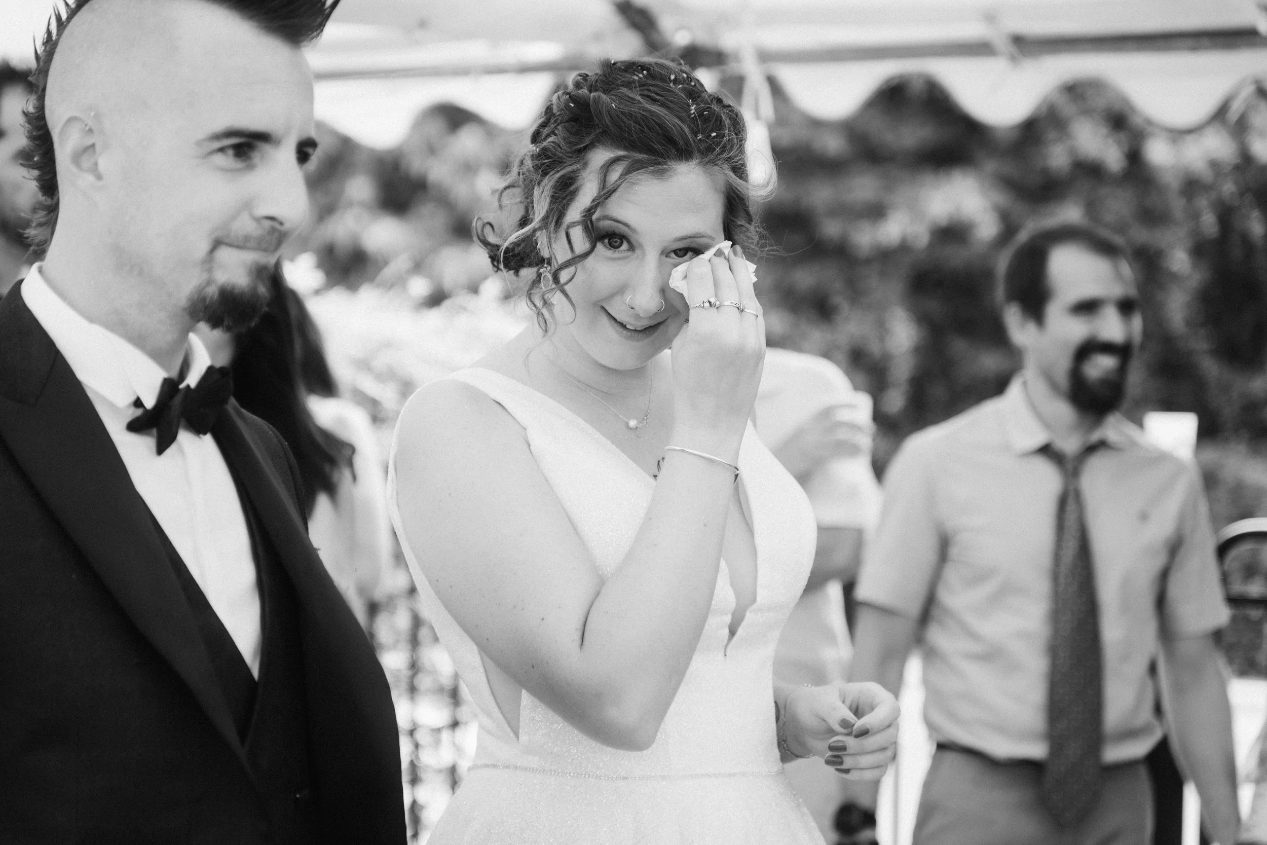 Une femme en robe blanche, à un mariage, sourit et pleure en se mouchant, entourée d'autres personnes, notamment un homme en costume noir et une autre en chemise avec nœud papillon, lors d'une cérémonie en extérieur.