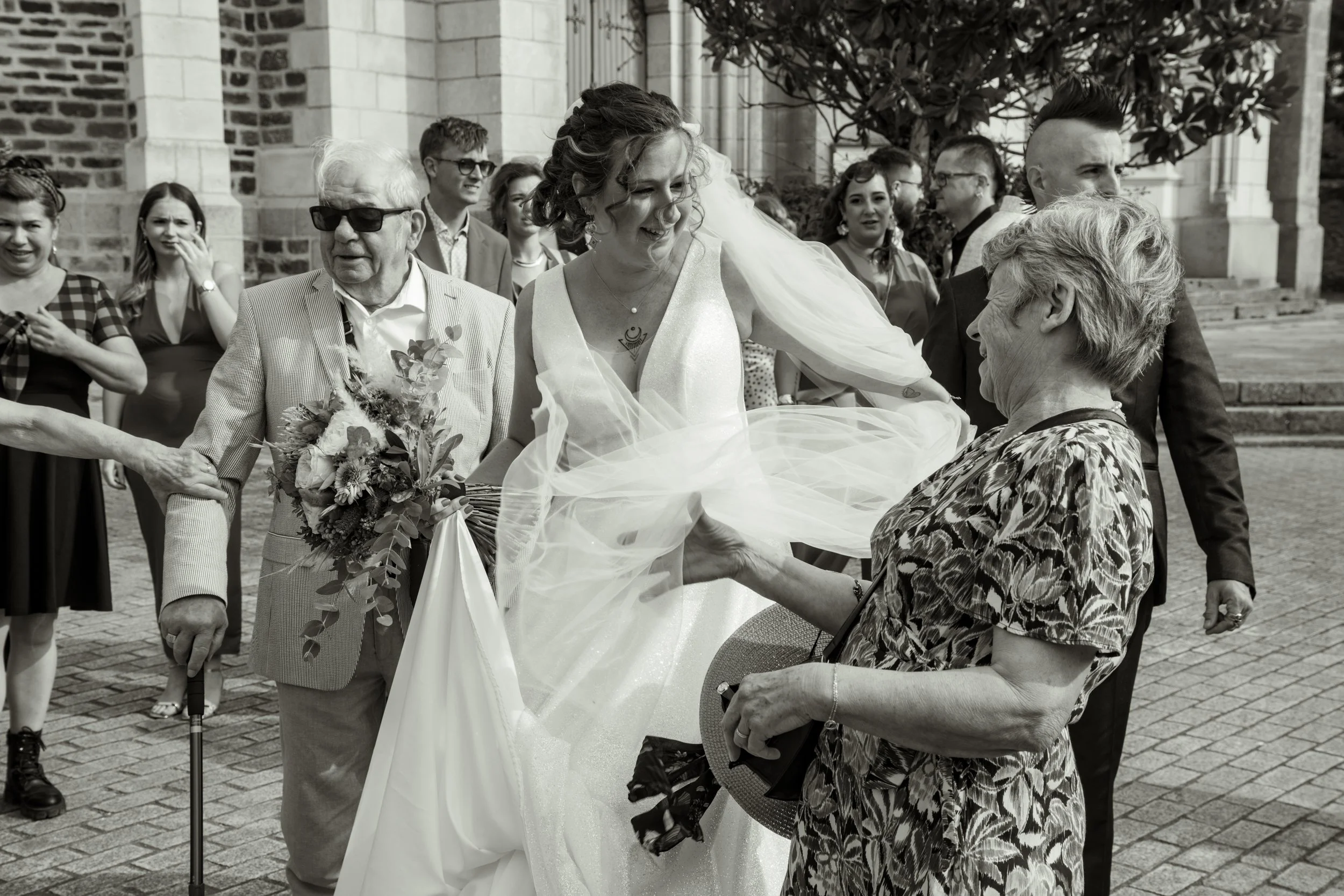 Une femme en robe de mariage sourit alors qu'elle est aidée à descendre d'une chaise par une femme âgée lors d'un mariage en extérieur, avec d'autres invités en arrière-plan.