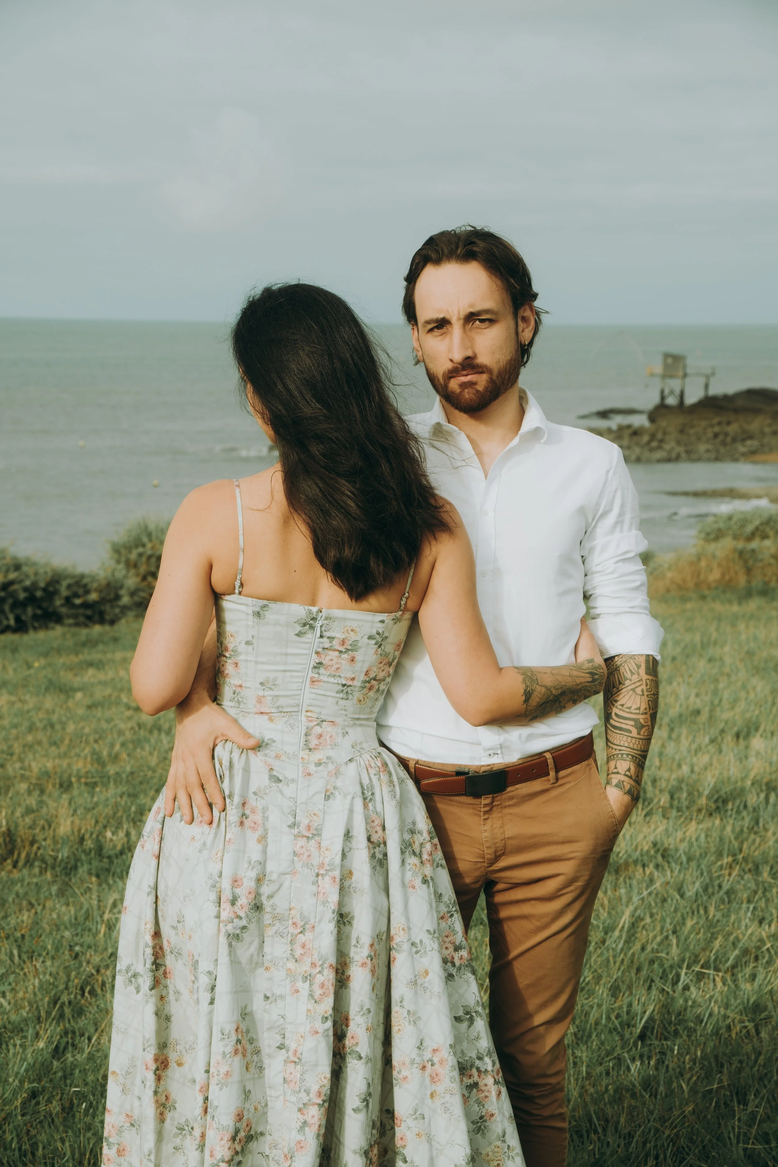Un homme et une femme debout dans une zone herbeuse près de la mer, la femme fait face à l'homme, qui regarde directement la caméra, dans un décor côtier avec une structure de cabane en arrière-plan.