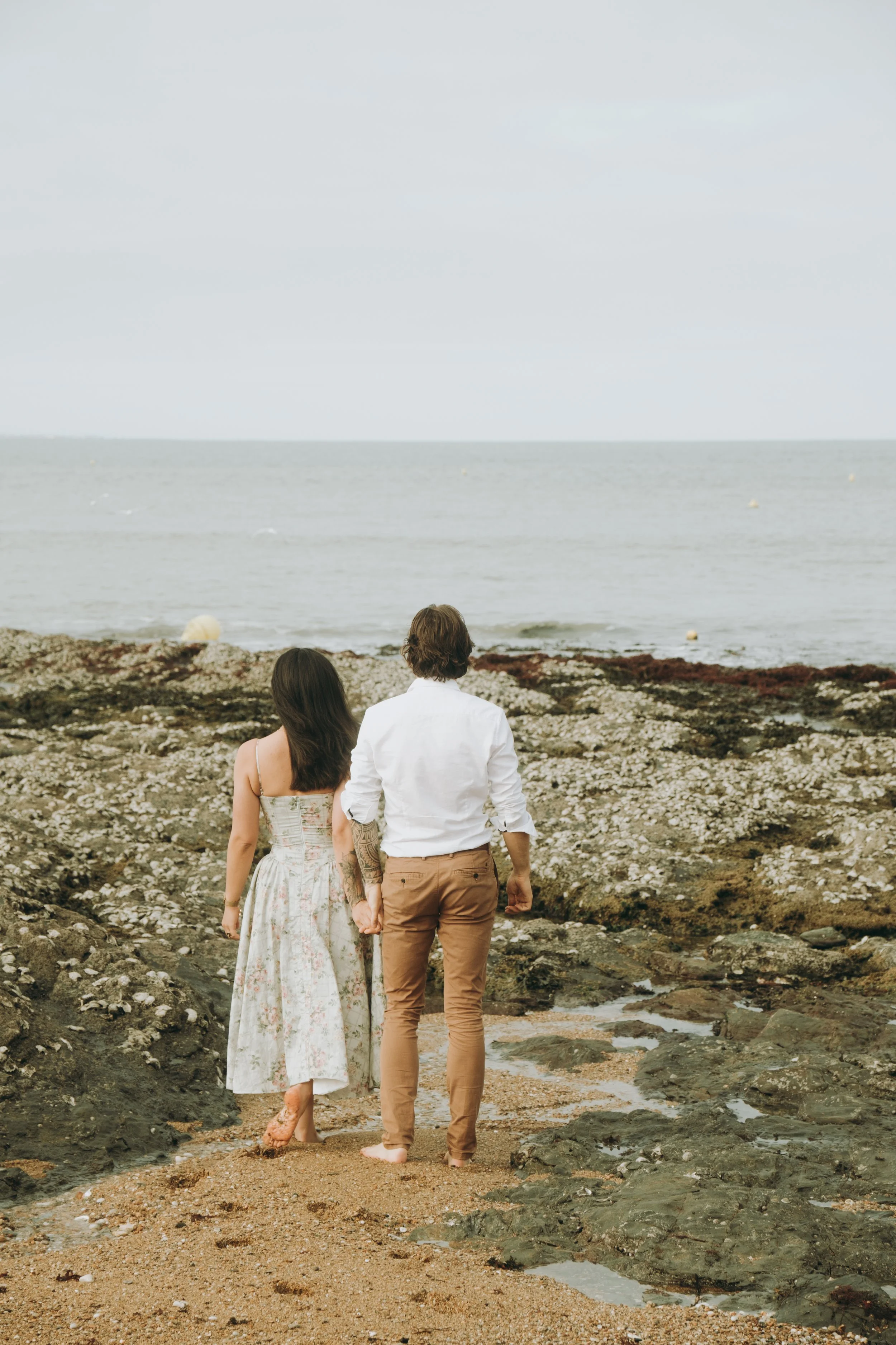 Un couple marche pieds nus sur la plage rocheuse en direction de la mer, vue de dos.