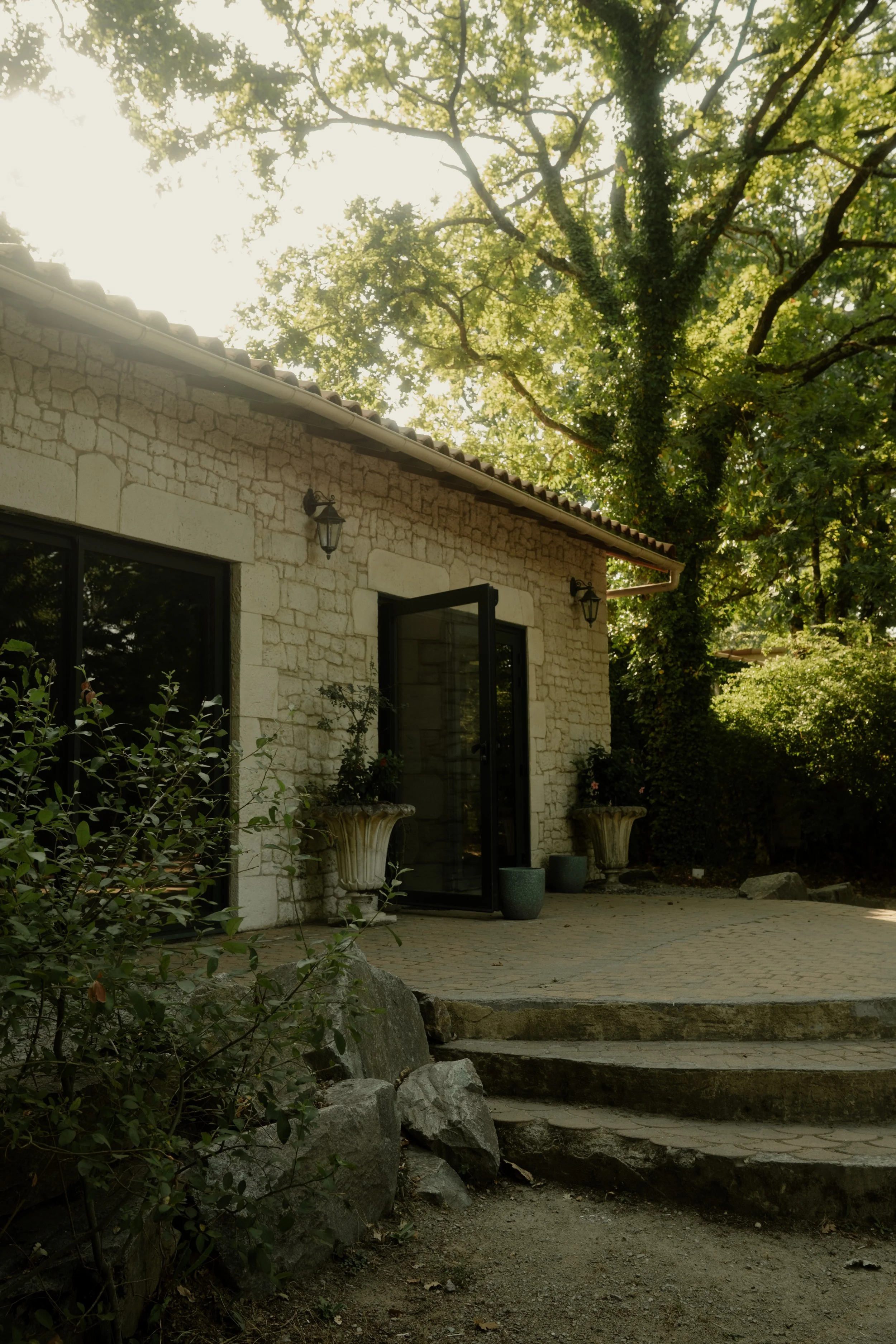 maison en pierre, entrée avec portes vitrées, plantes en pot, arbres environnants, escalier en pierre.