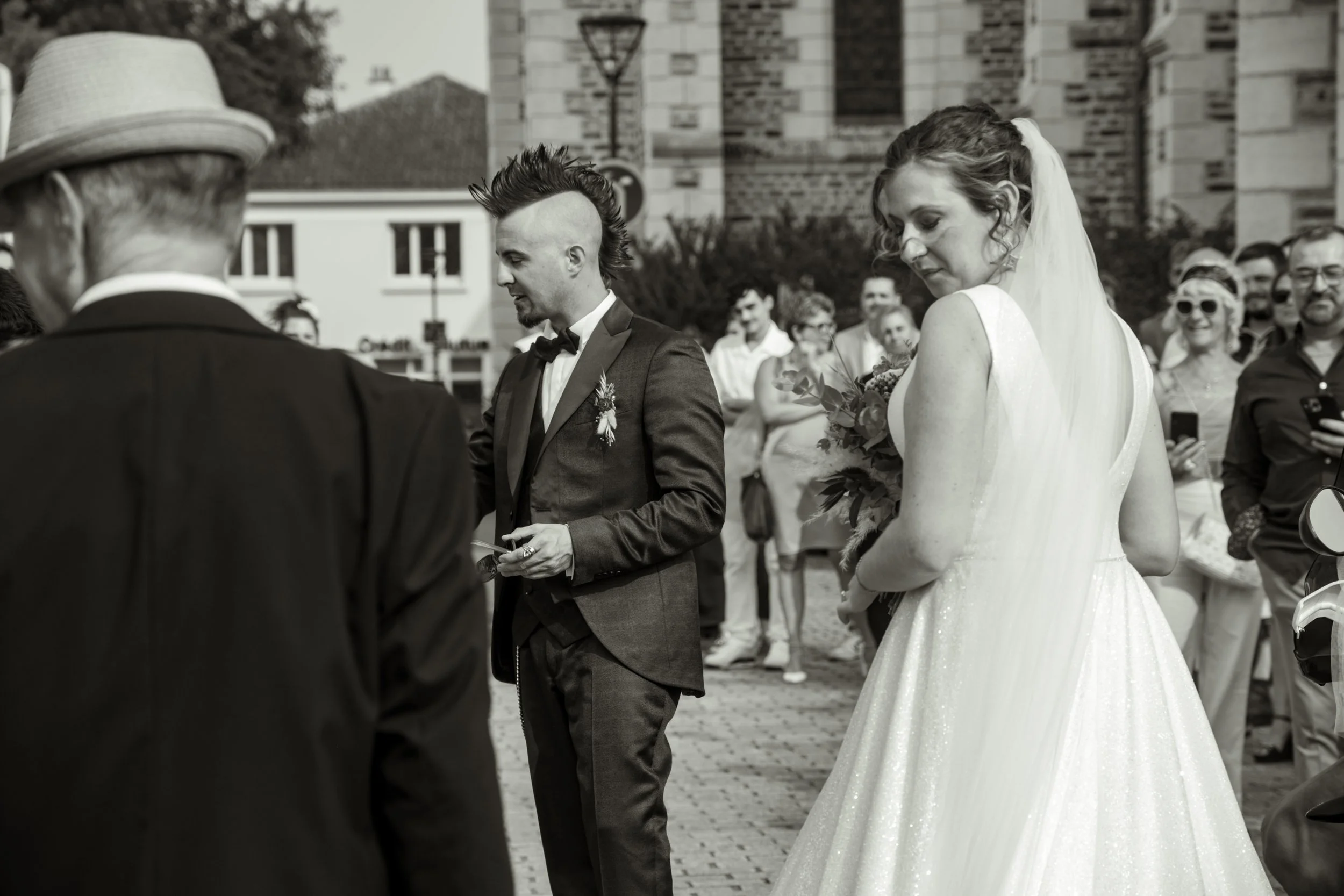 Un mariage en plein air avec un couple de mariés et des invités, scène en noir et blanc. La mariée porte une robe de mariage, le marié porte un costume avec un nœud papillon, et ils sont entourés de personnes souriantes.