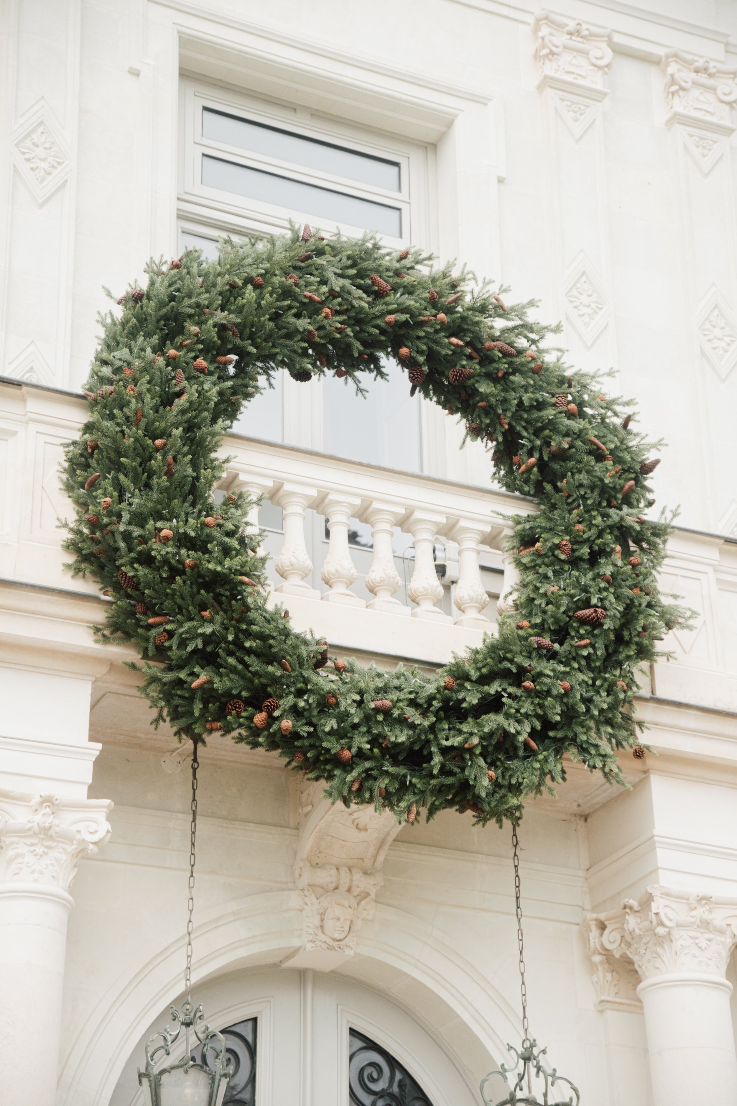 Une couronne de Noël faite de branches de sapin avec des cônes de pin suspendue sur une façade de bâtiment blanc avec une fenêtre et un balcon.