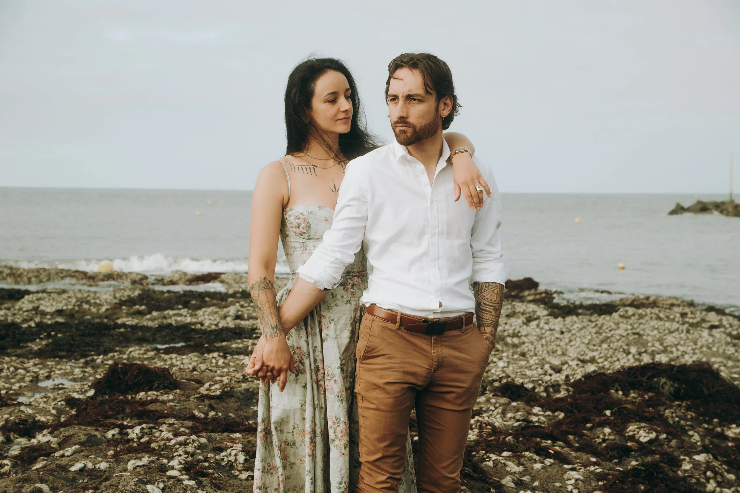 Un homme et une femme tenant la main sur une plage rocheuse, la femme repose sa main sur l'épaule de l'homme, tous deux regardent dans des directions différentes, arrière-plan de la mer et ciel nuageux.