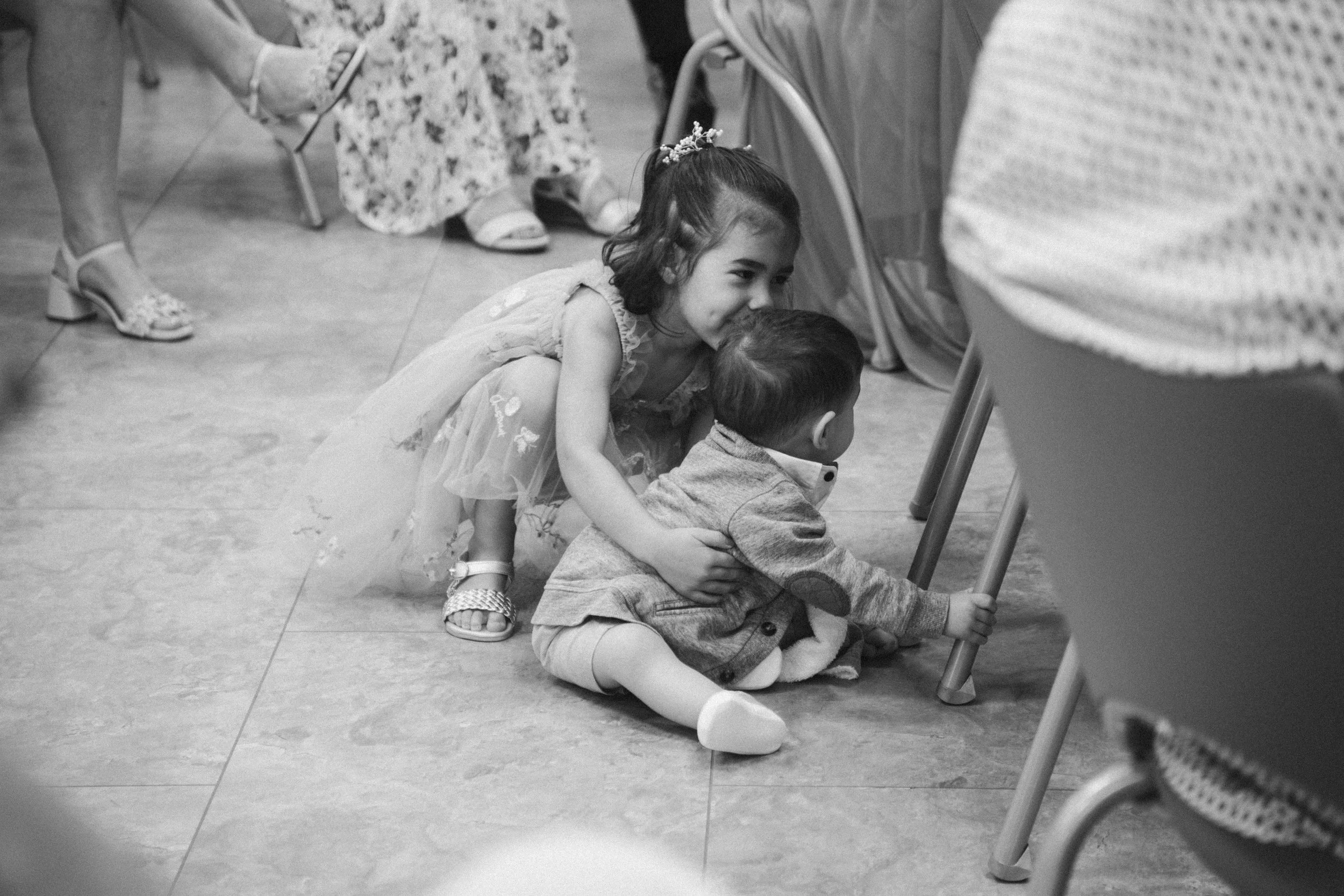 Deux enfants jouent par terre lors d'une fête ou d'un rassemblement, la fille semble tirer le garçon par la main. Autour, il y a des adultes assis et des chaises.
