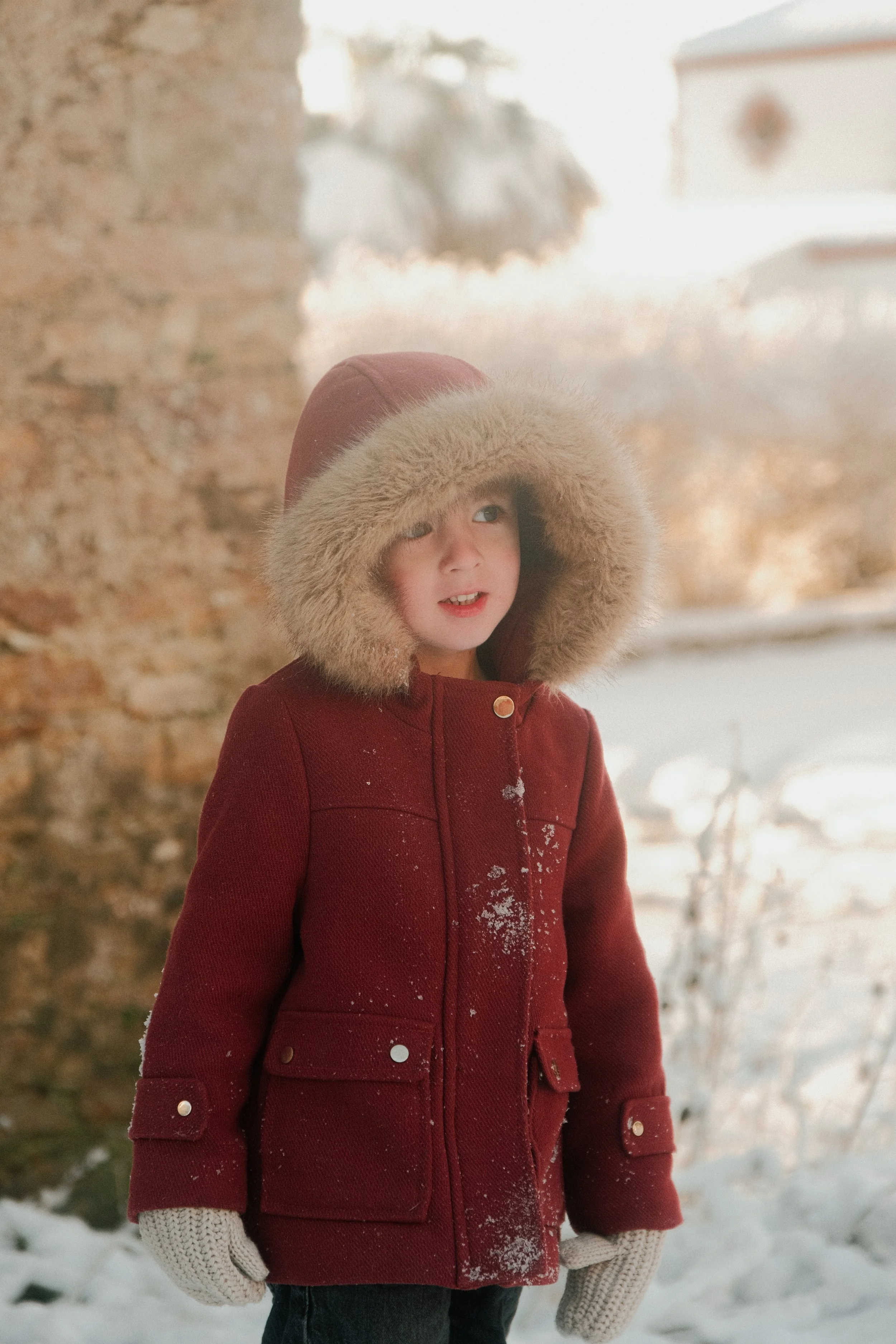 Jeune enfant habillé d'un manteau rouge avec une capuche en fourrure, portant des gants, dans un décor enneigé.