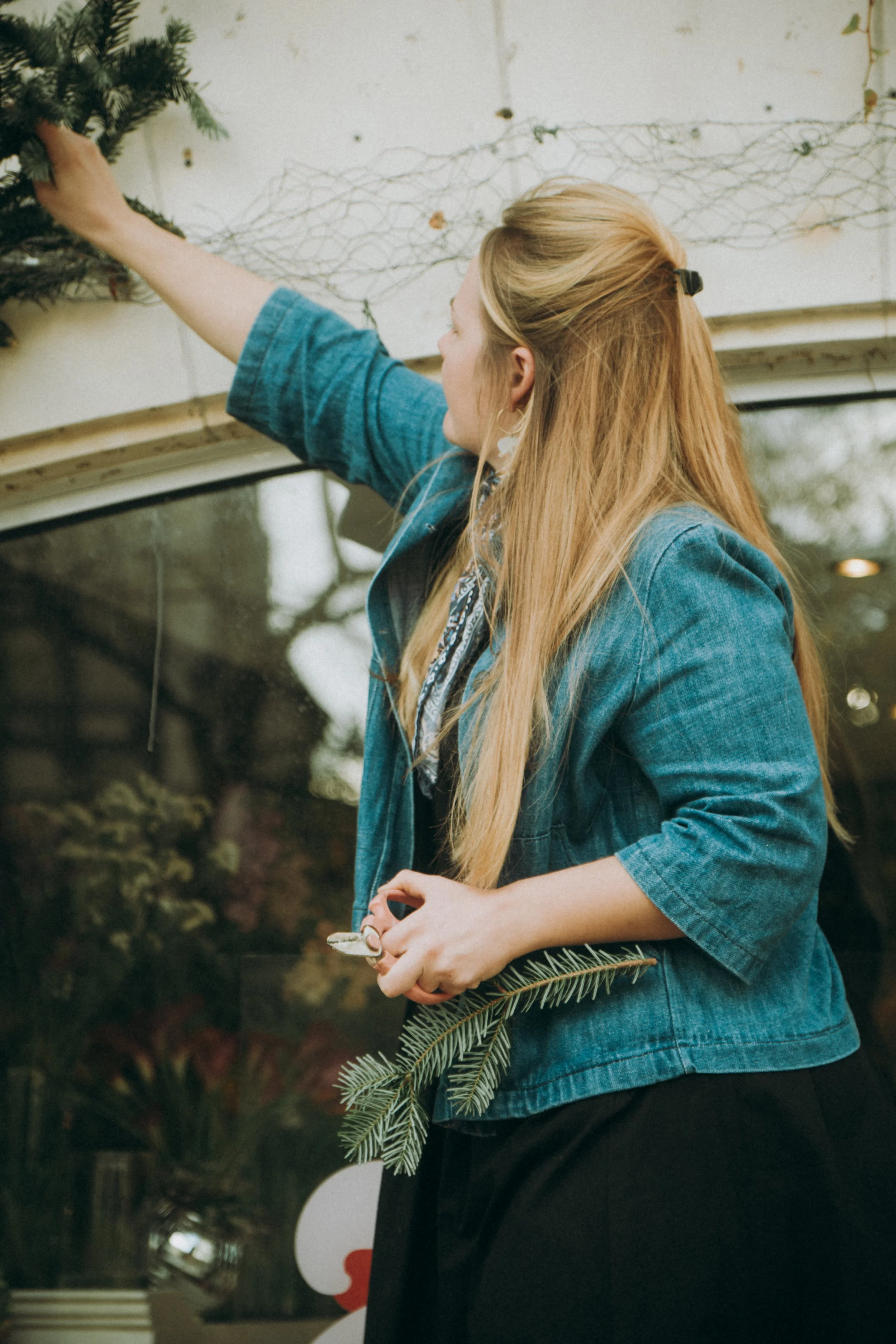 Une femme à cheveux longs, portant une veste en jean, décore une fenêtre avec une branche de sapin pour Noël.